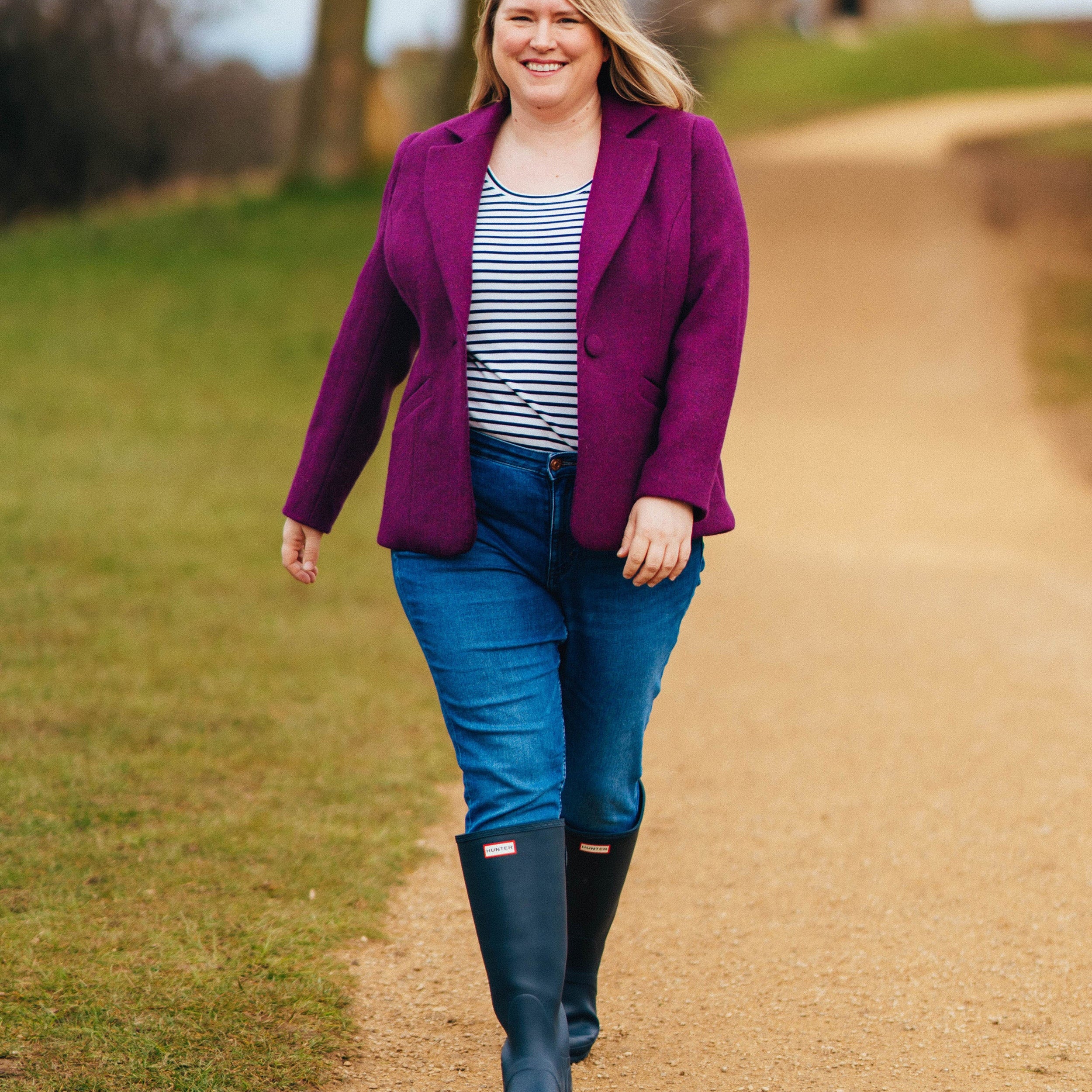 Woman in a purple coat, striped shirt, and blue jeans walking on a path outdoors.. This blazer is the Auburn Blazer sewing pattern from Cashmerette.
