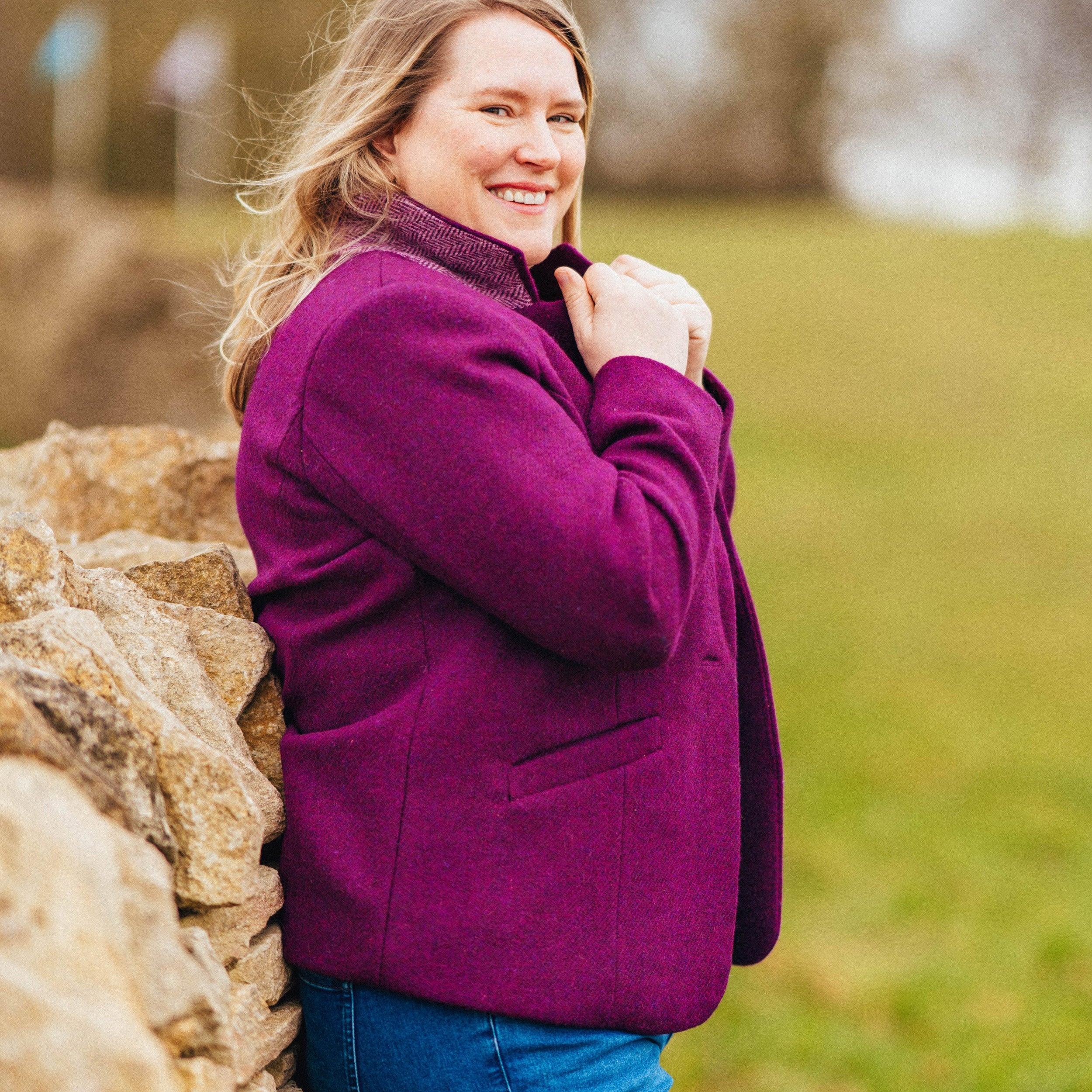 Woman in a purple coat standing next to a stone wall with a blurred natural background. This blazer is the Auburn Blazer sewing pattern from Cashmerette.