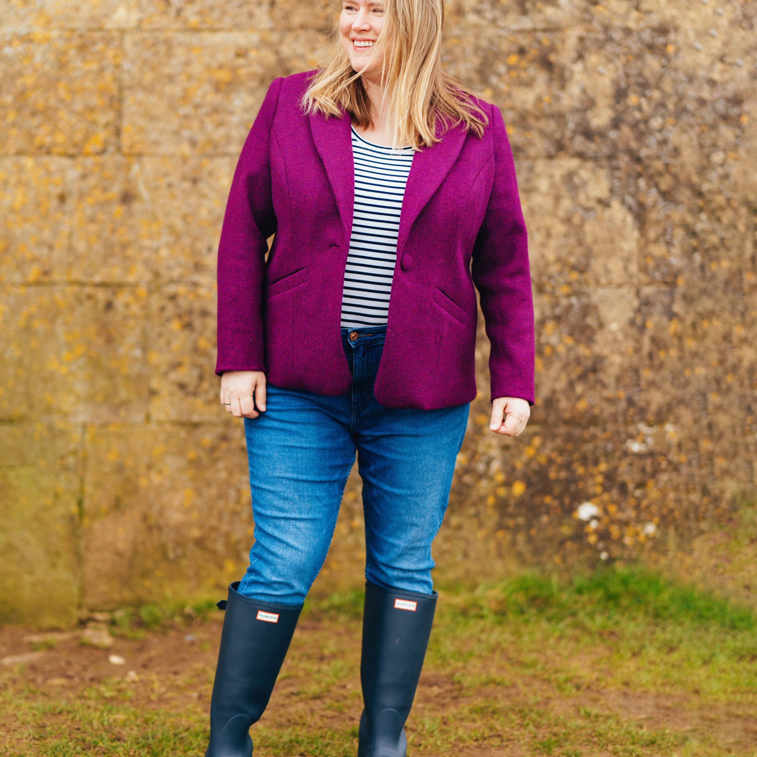 Woman wearing a purple jacket, striped shirt, blue jeans, and navy boots standing in front of a stone wall. This blazer is the Auburn Blazer sewing pattern from Cashmerette.