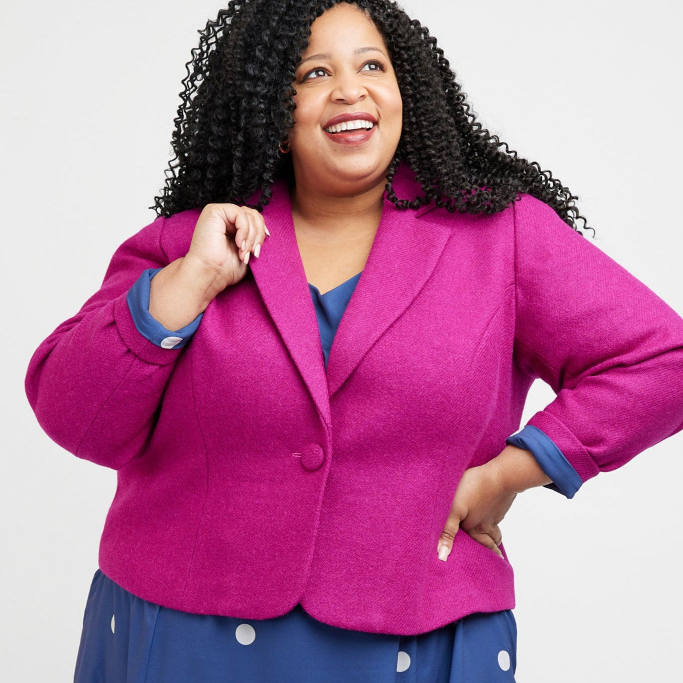 Woman wearing a bright pink blazer over a blue dress with white polka dots on a white background. This blazer is the Auburn Blazer sewing pattern from Cashmerette.