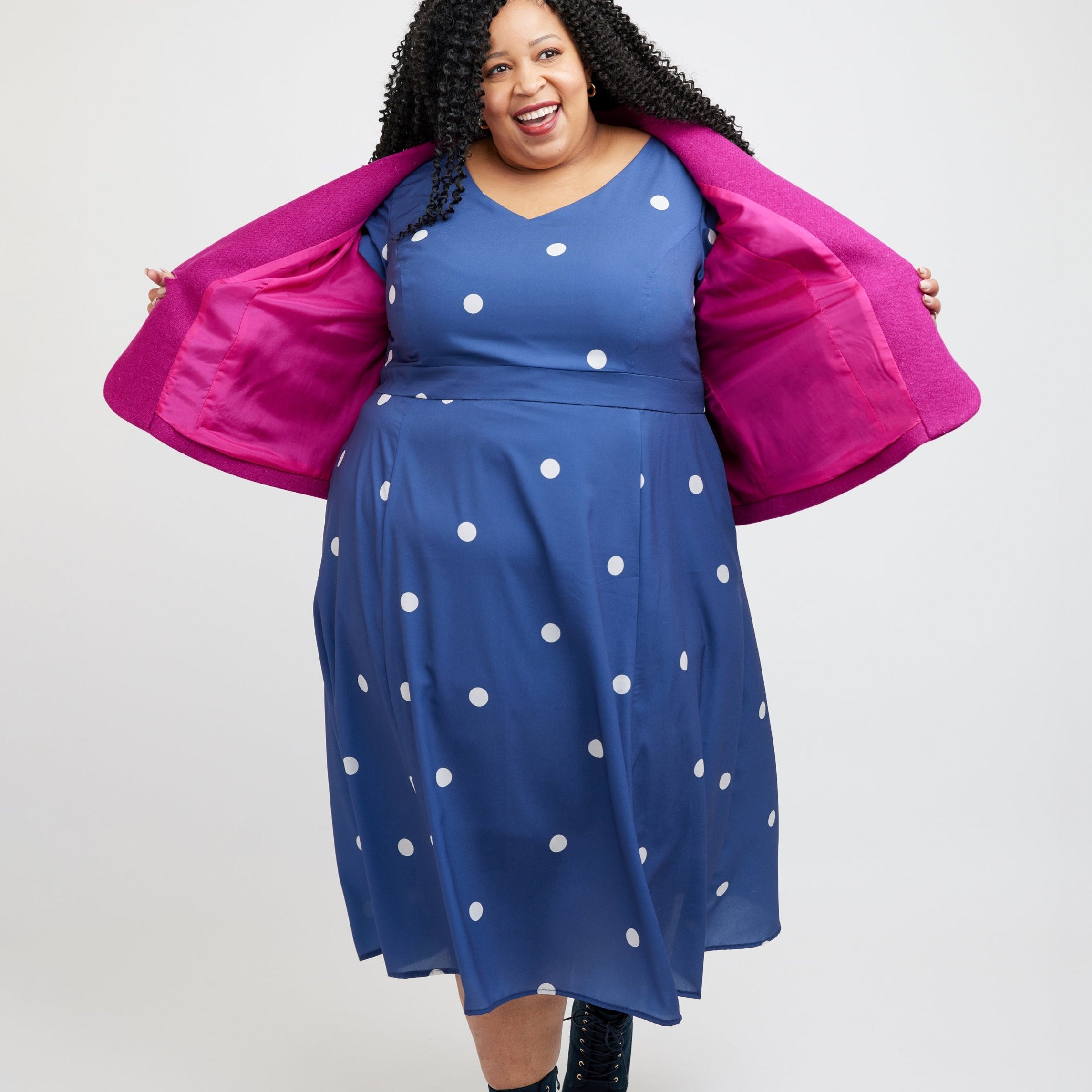 Woman wearing a blue dress with white polka dots and a pink jacket on a white background. This blazer is the Auburn Blazer sewing pattern from Cashmerette.
