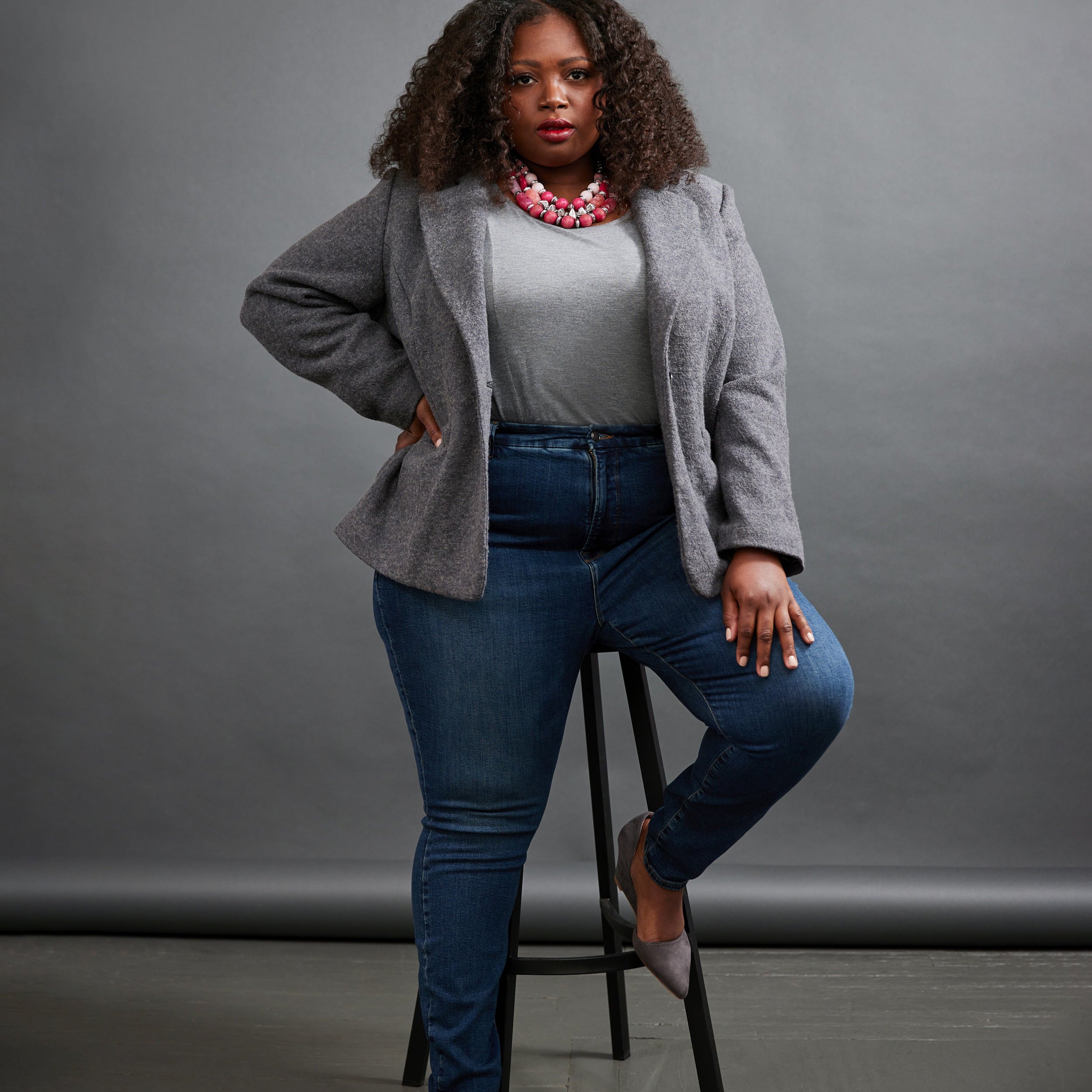 Woman sitting on a stool wearing a gray cardigan, white shirt, blue jeans, and gray shoes against a gray background. This blazer is the Auburn Blazer sewing pattern from Cashmerette.