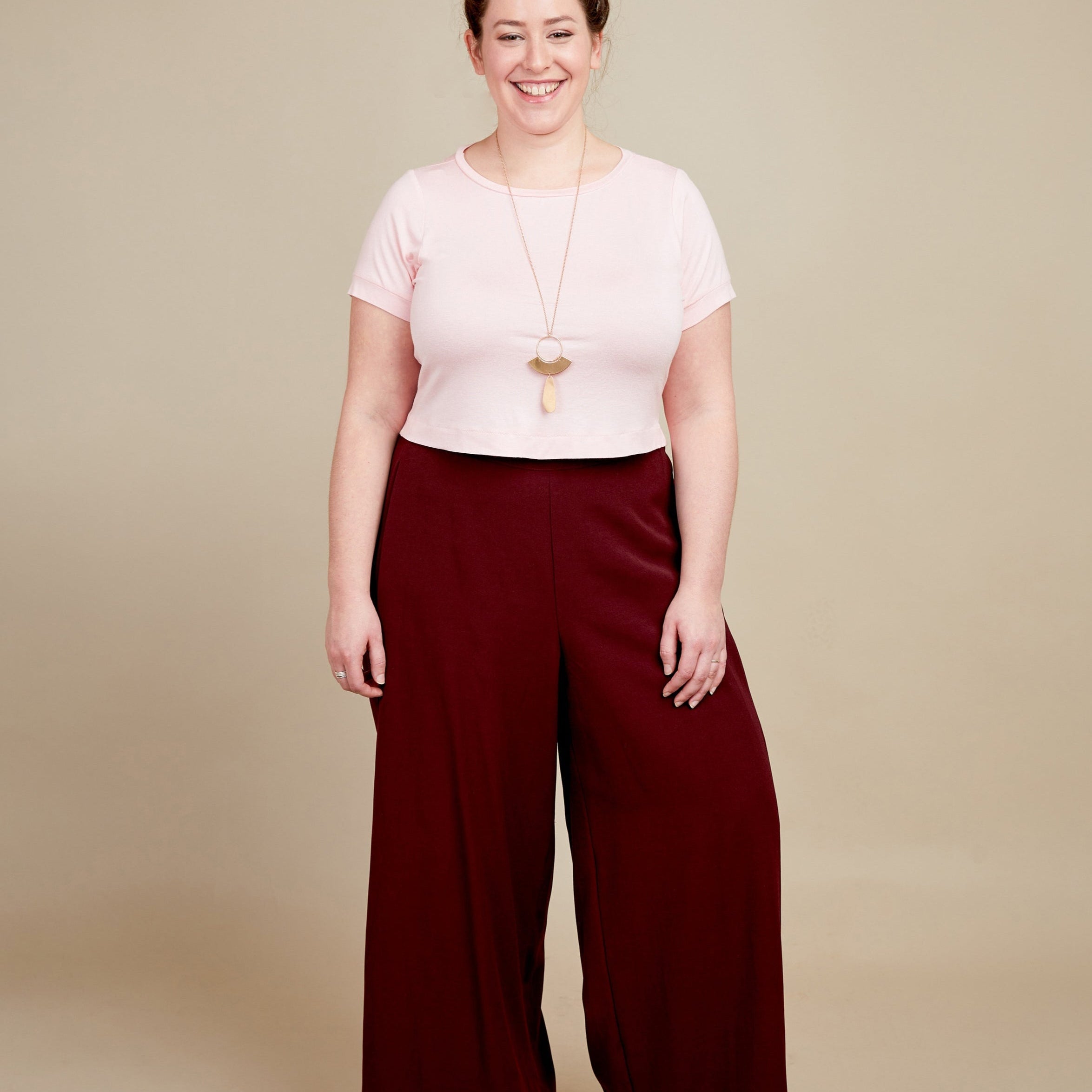 Woman wearing a pink top and burgundy wide-leg pants on a beige background. This t-shirt is the Concord T-Shirt sewing pattern from Cashmerette.