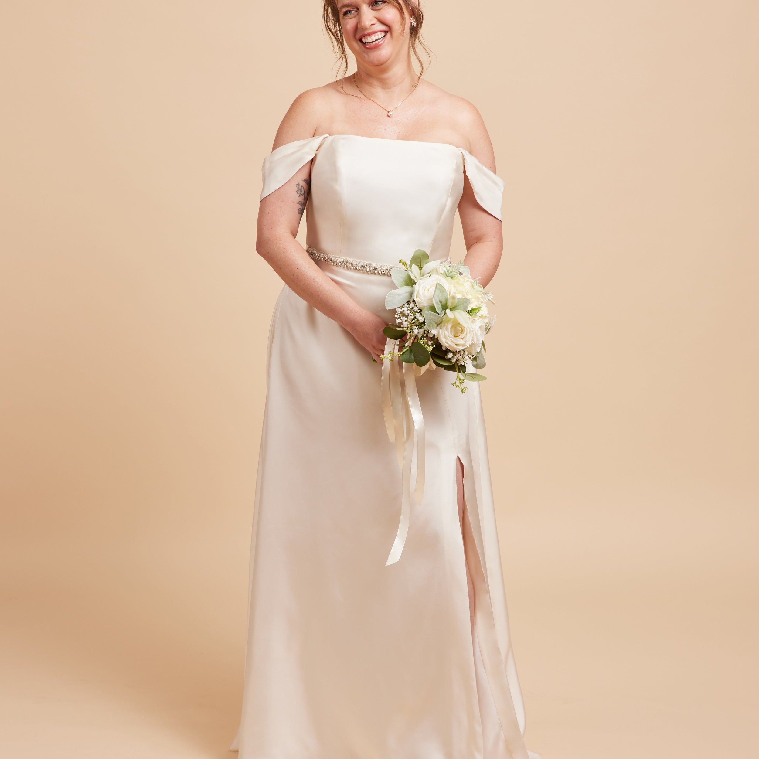 Woman in a white off-shoulder wedding dress holding a bouquet on a beige background. This dress is the Ellington Strapless Dress sewing pattern from Cashmerette.