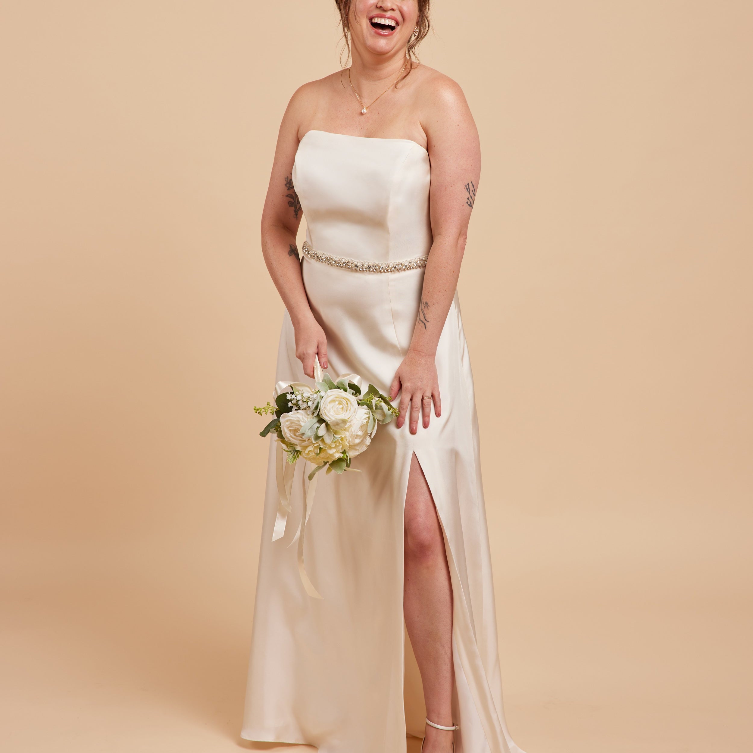Woman in a strapless white wedding dress with a high slit, holding a bouquet on a beige background. This dress is the Ellington Strapless Dress sewing pattern from Cashmerette.