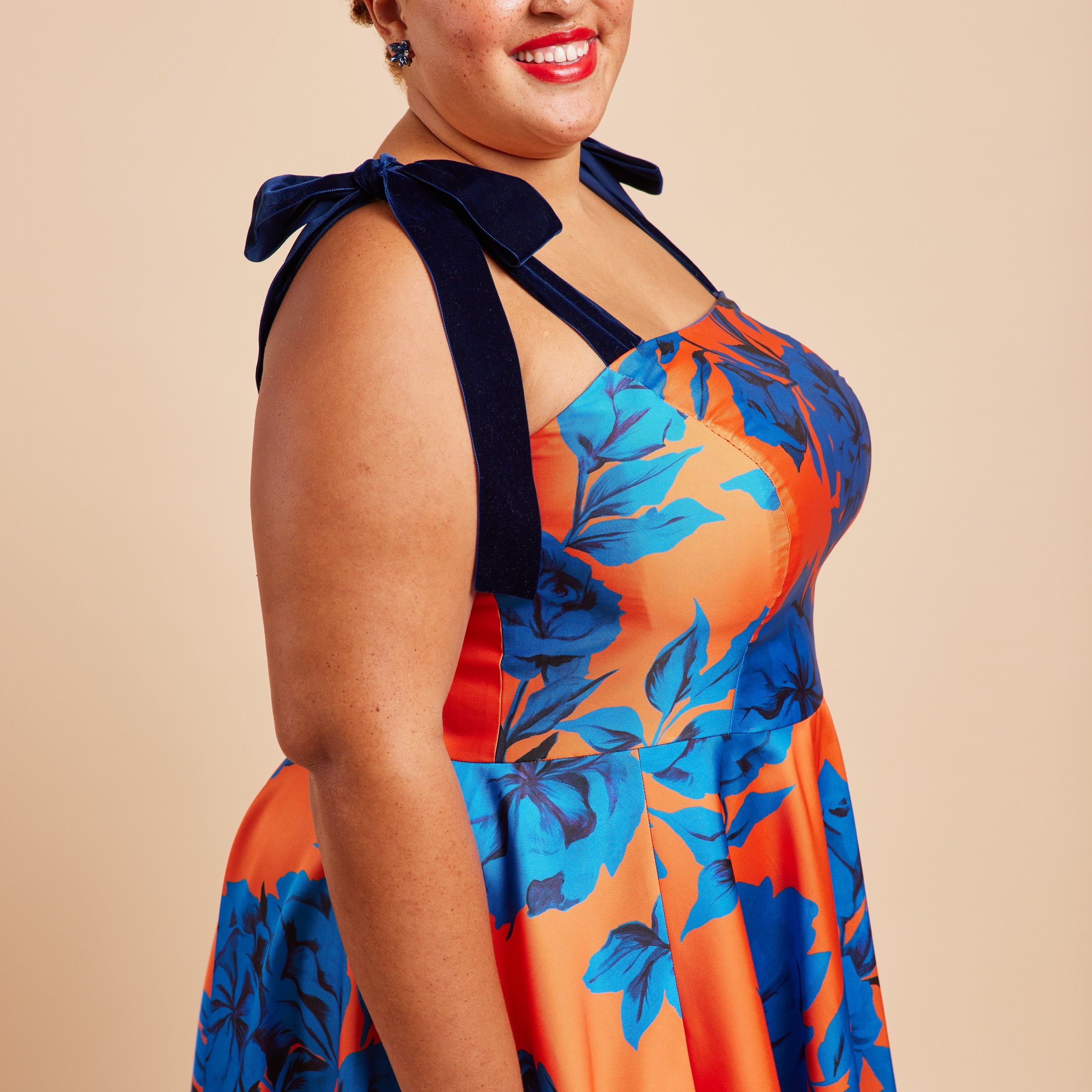 Woman wearing a colorful floral dress with a bow detail on a beige background. This dress is the Ellington Strapless Dress sewing pattern from Cashmerette.