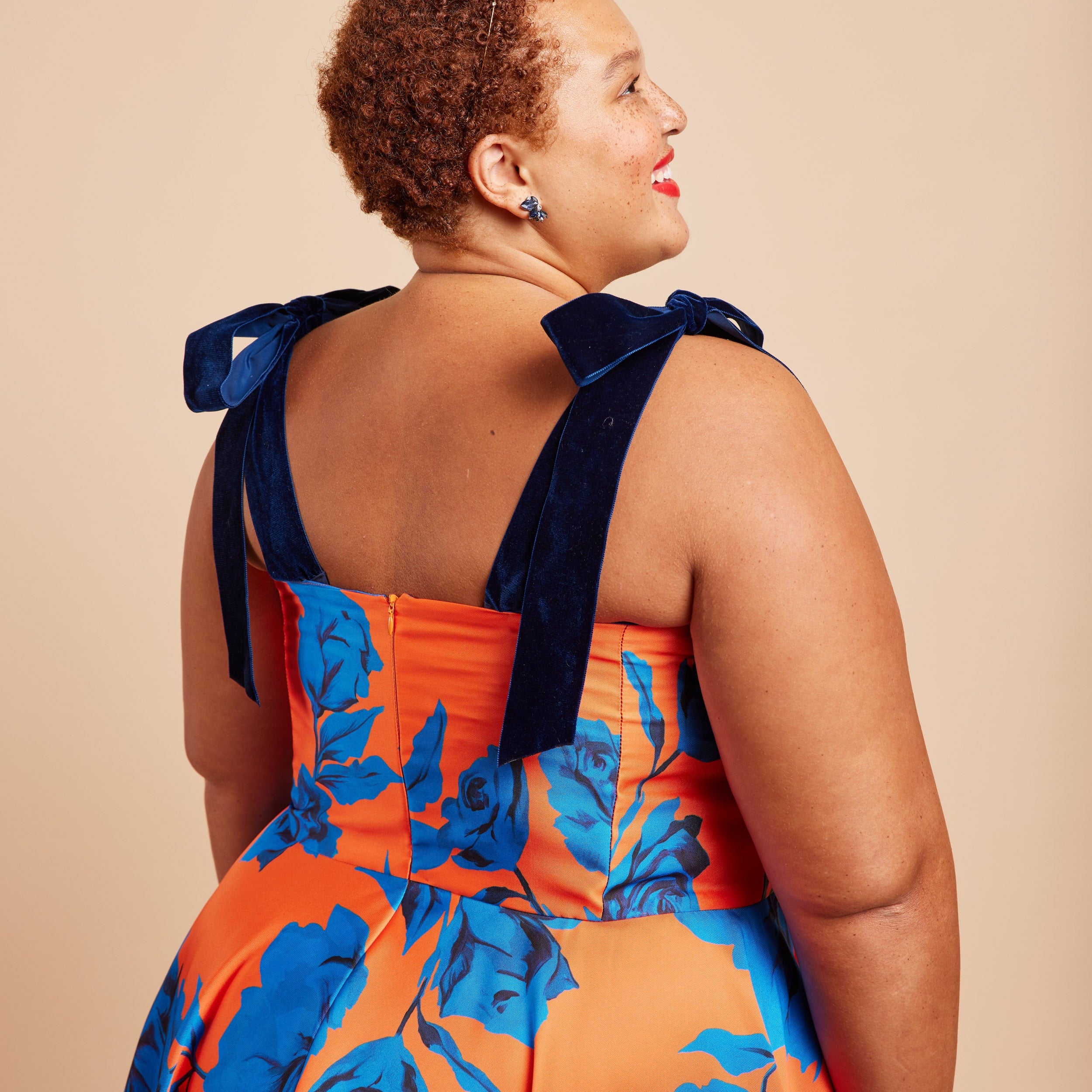 Woman wearing an orange and blue floral dress with bow straps on a beige background. This dress is the Ellington Strapless Dress sewing pattern from Cashmerette.