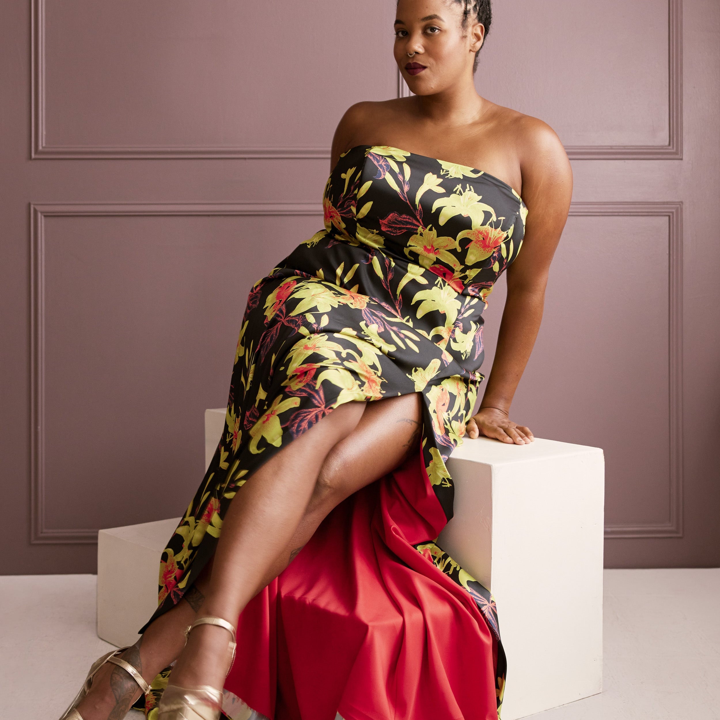Woman in a floral strapless dress sitting on a white cube against a purple wall. This dress is the Ellington Strapless Dress sewing pattern from Cashmerette.