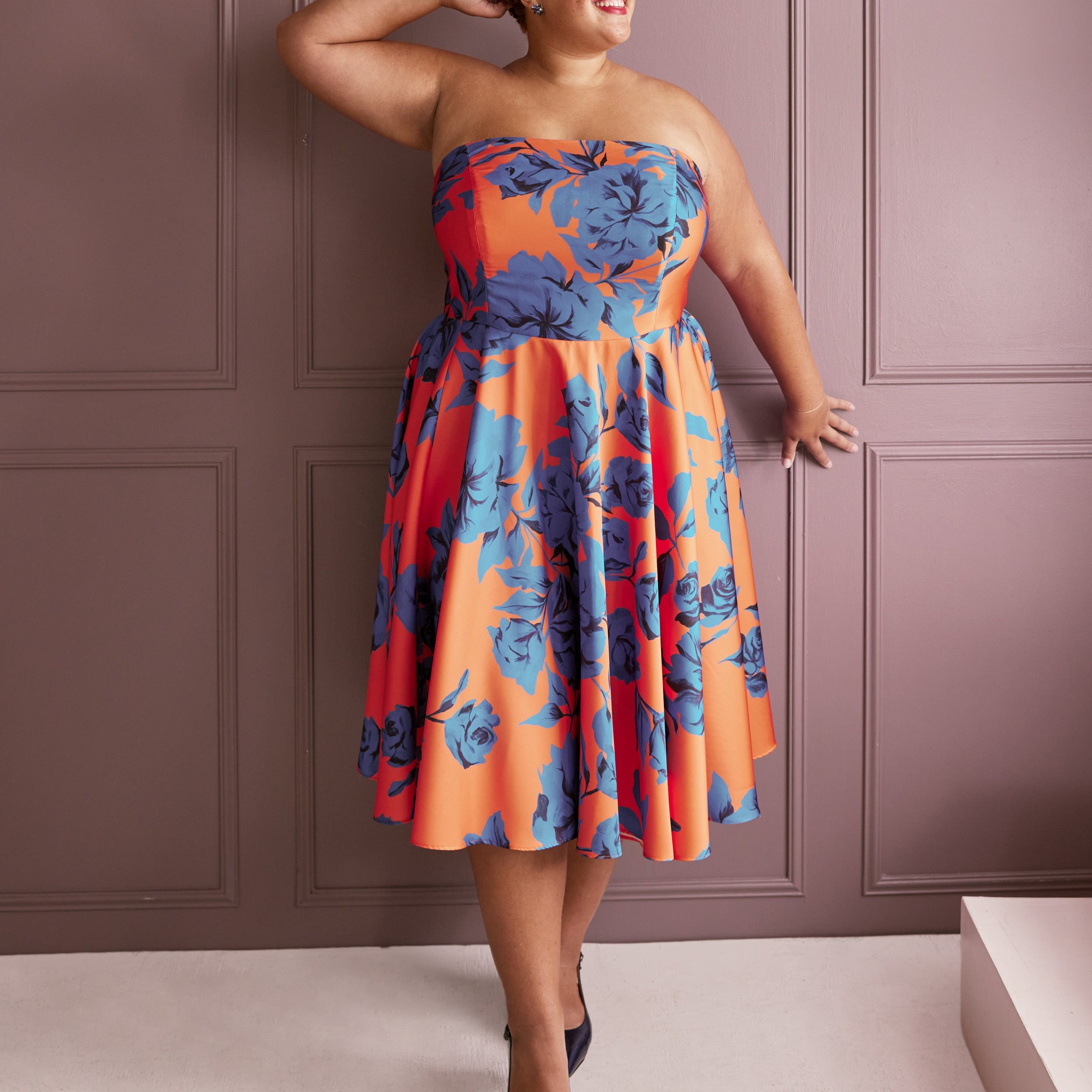 Woman wearing a strapless dress with blue floral pattern against a pink paneled wall. This dress is the Ellington Strapless Dress sewing pattern from Cashmerette.