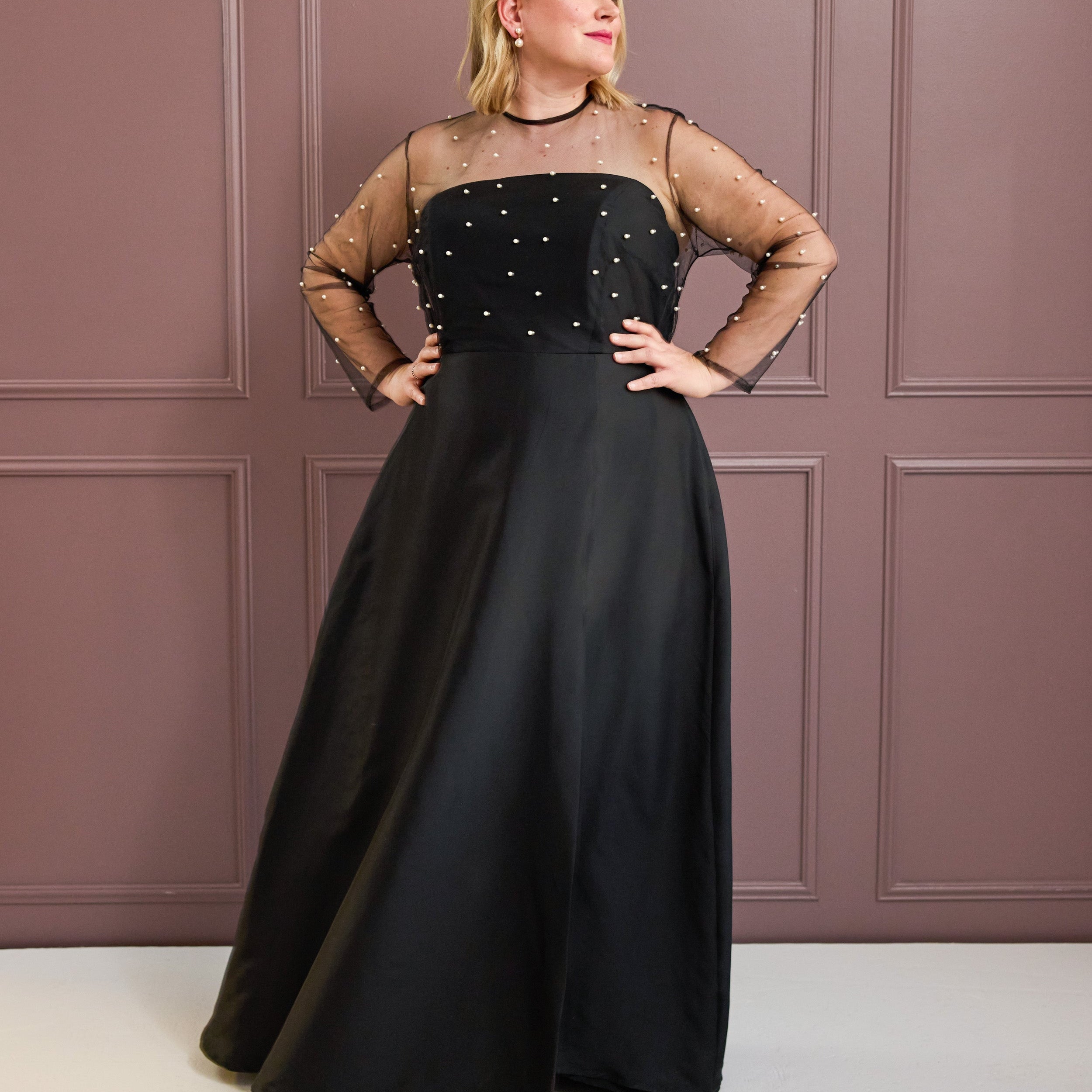 Woman wearing a black evening gown with sheer sleeves against a pink paneled wall. This is the Ellington Dress sewing pattern from Cashmerette.