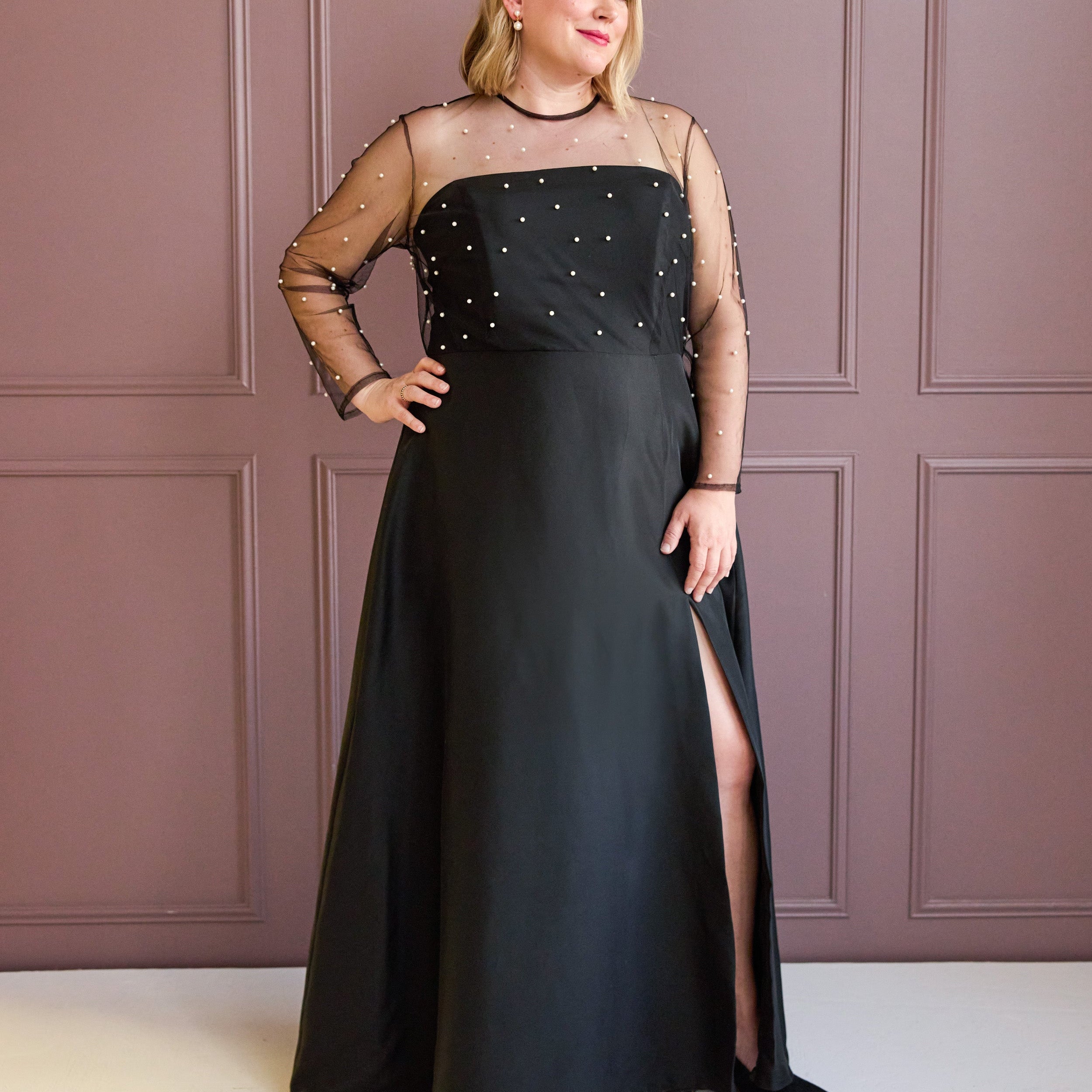 Woman wearing a black evening gown with sheer sleeves against a pink paneled wall. This is the Ellington Dress sewing pattern from Cashmerette.