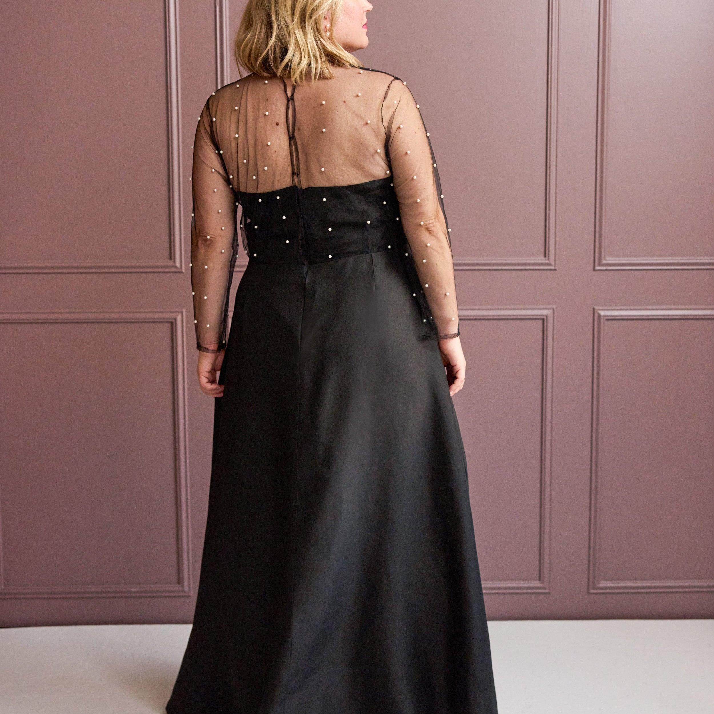 Woman wearing a black evening gown with sheer sleeves against a pink paneled wall. This is the Ellington Dress sewing pattern from Cashmerette.