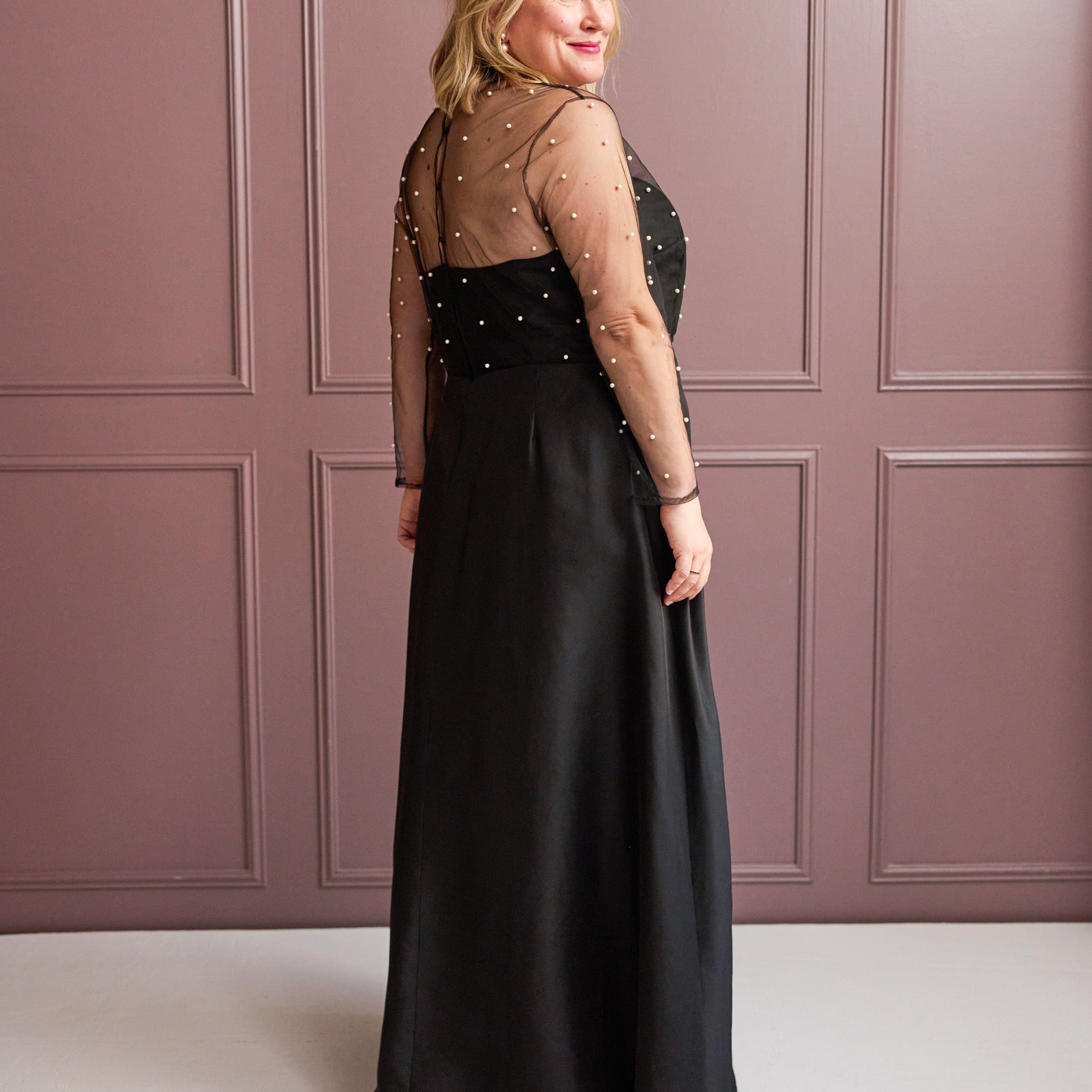 Woman wearing a black evening gown with sheer sleeves against a pink paneled wall. This is the Ellington Dress sewing pattern from Cashmerette.