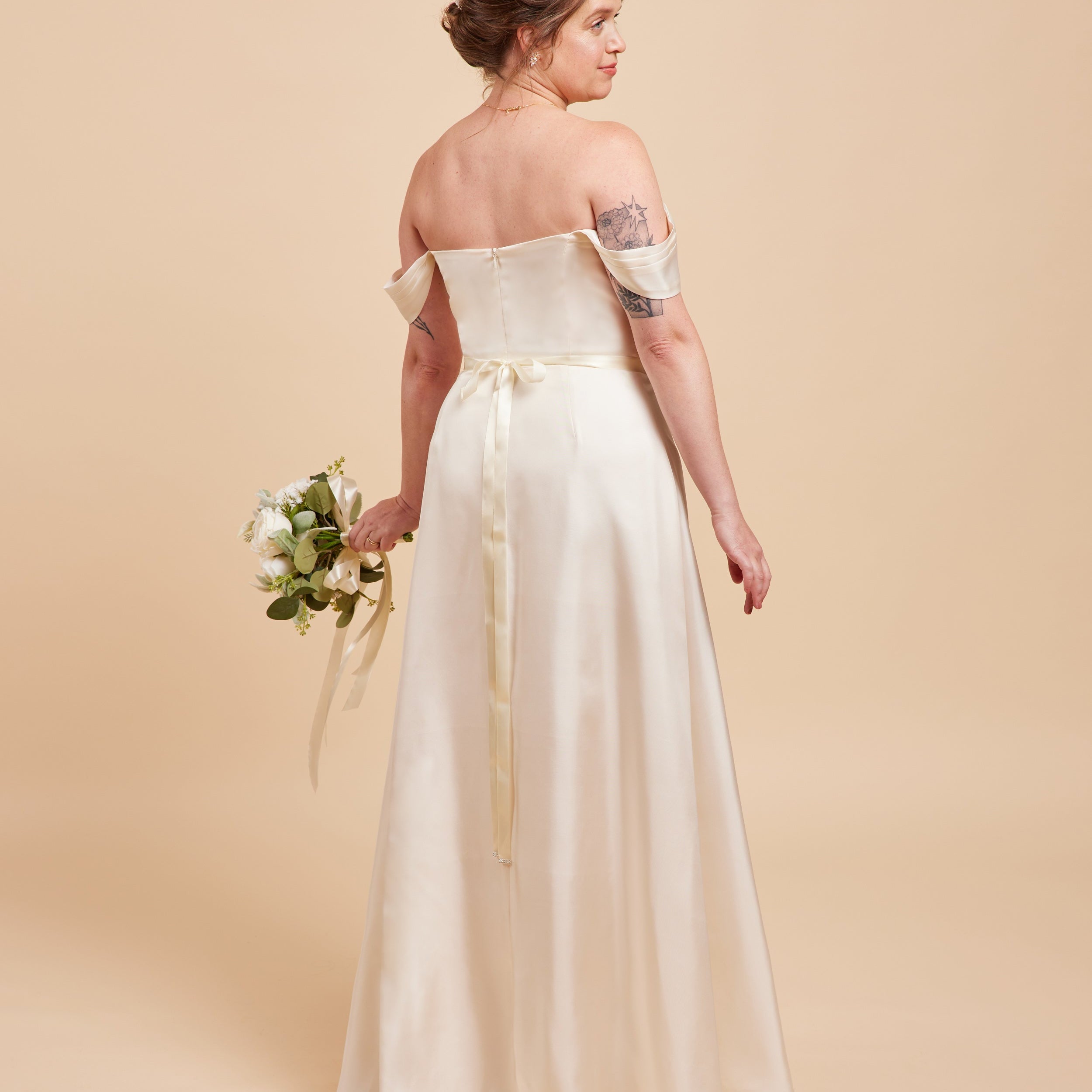 Woman in a white off-shoulder wedding dress holding a bouquet on a beige background. This dress is the Ellington Strapless Dress sewing pattern from Cashmerette.