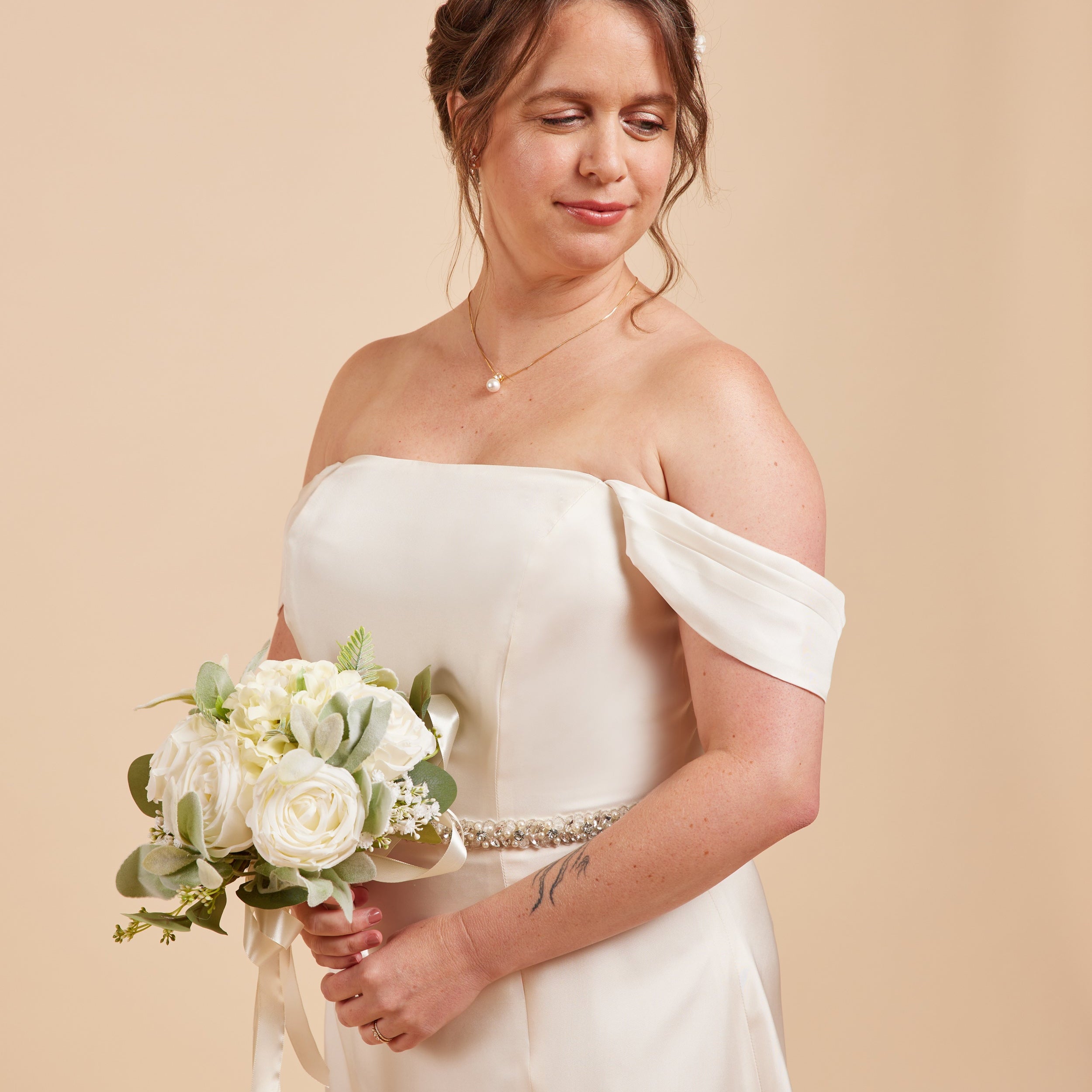 Woman in a white off-shoulder dress holding a bouquet against a beige background. This dress is the Ellington Strapless Dress sewing pattern from Cashmerette.