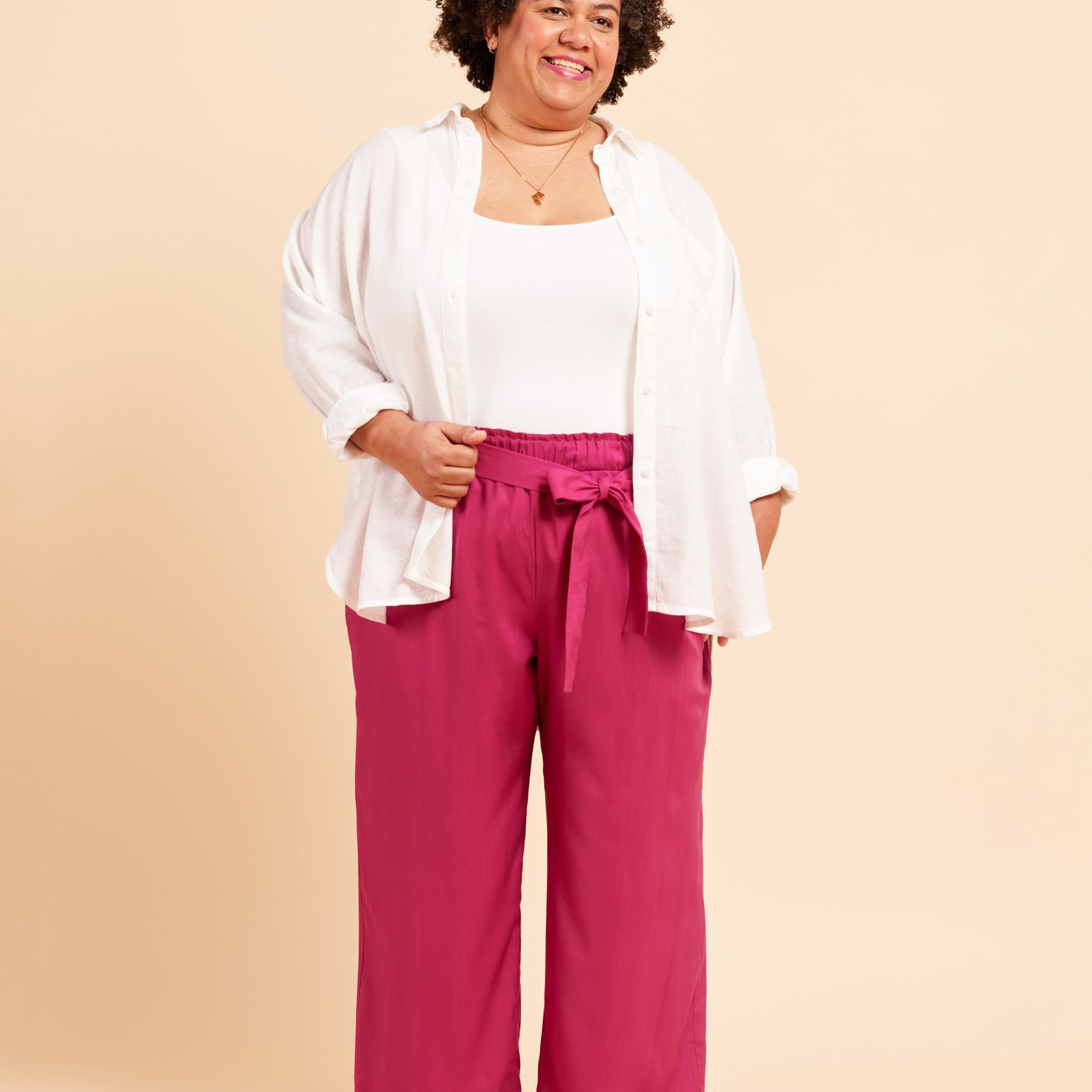 Woman wearing a white top and pink pants against a beige background. These pants are the Everett Pants & Shorts sewing pattern from Cashmerette.