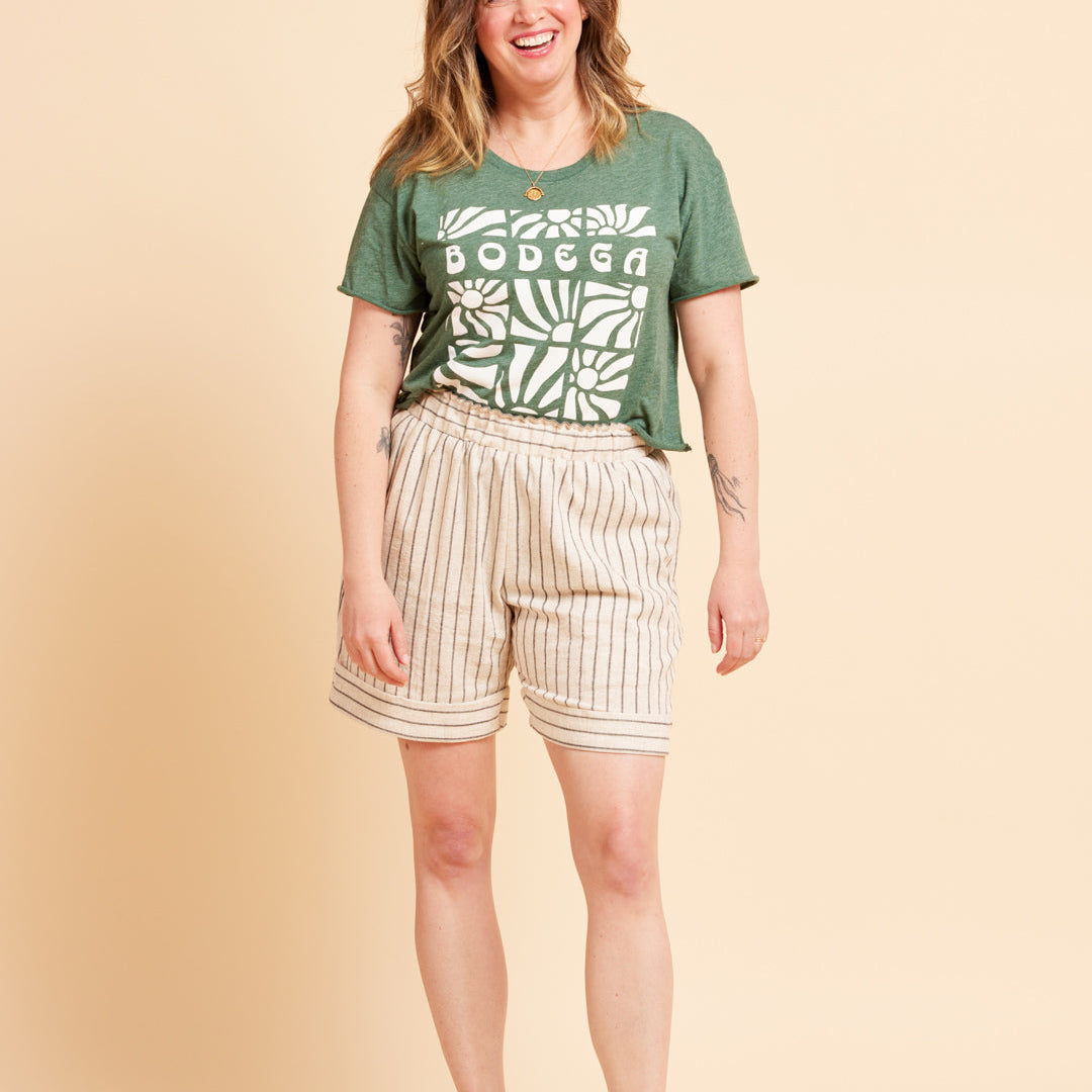 Woman wearing a green t-shirt with 'Bodega' print and beige shorts on a beige background. These shorts are the Everett Pants & Shorts sewing pattern from Cashmerette.