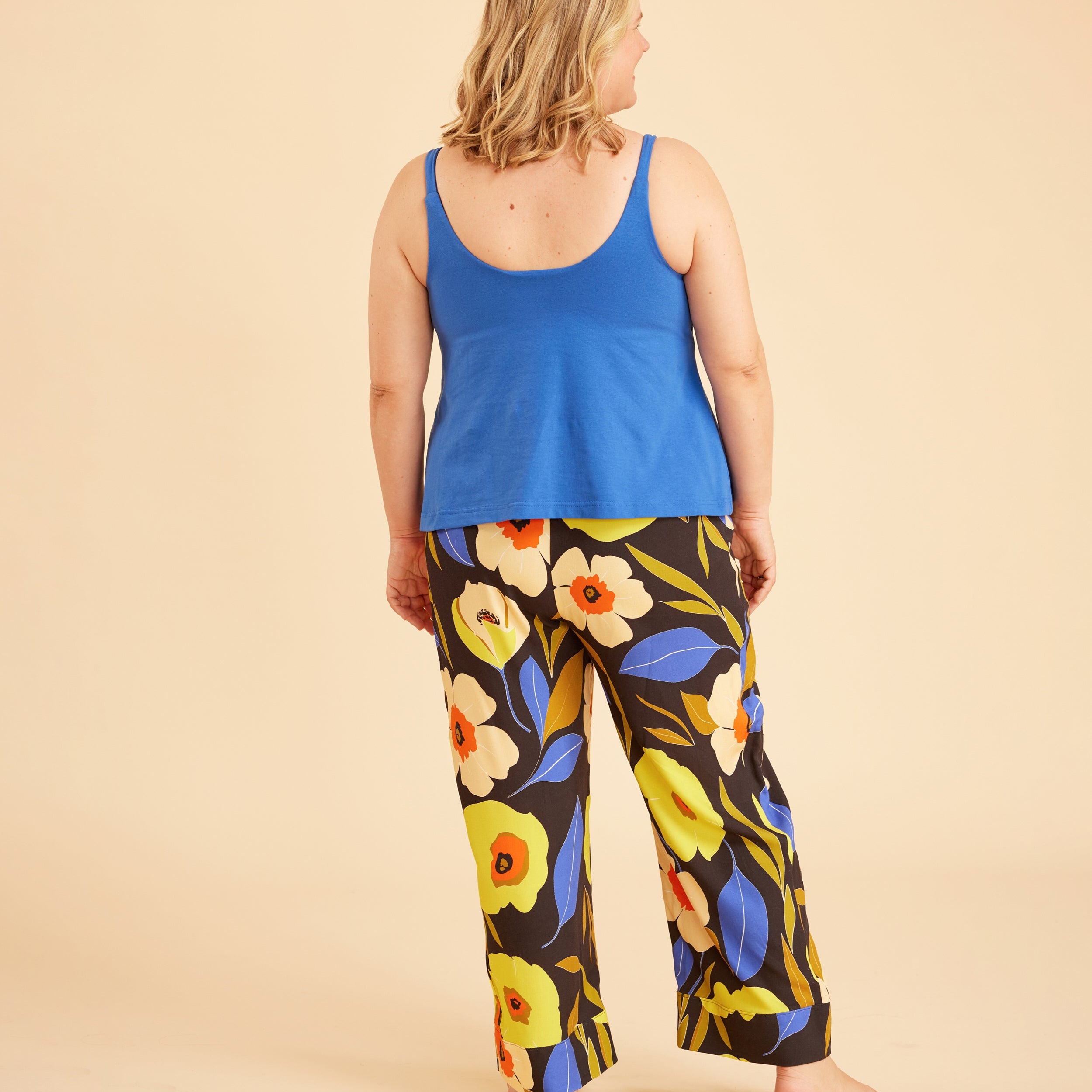 Woman wearing a blue tank top and colorful floral pants on a beige background. This is the Fiske Sleepwear Set sewing pattern from Cashmerette.
