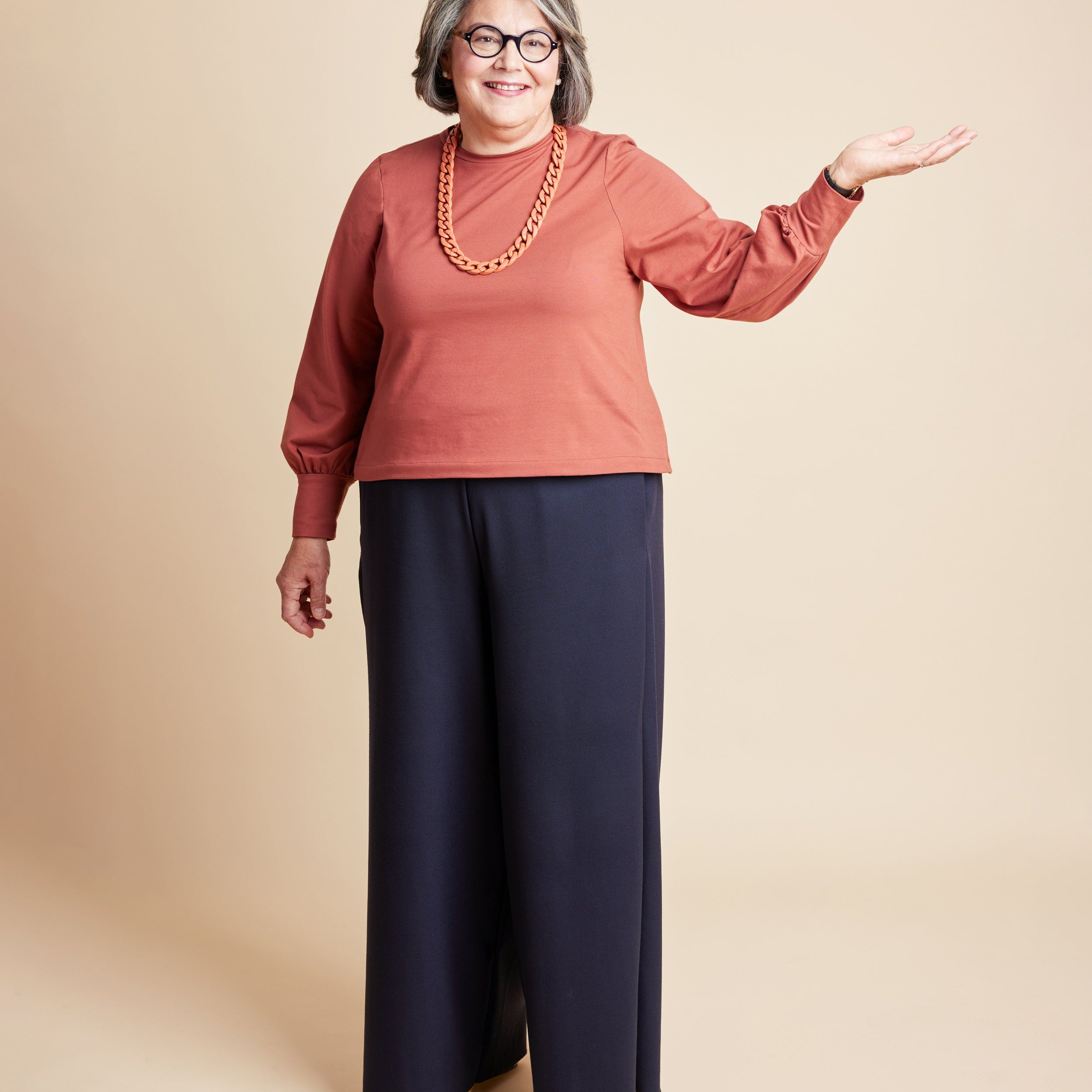 Woman wearing a rust-colored sweater and navy pants on a beige background. This is the Grafton Dress, Top & Skirt sewing pattern from Cashmerette.