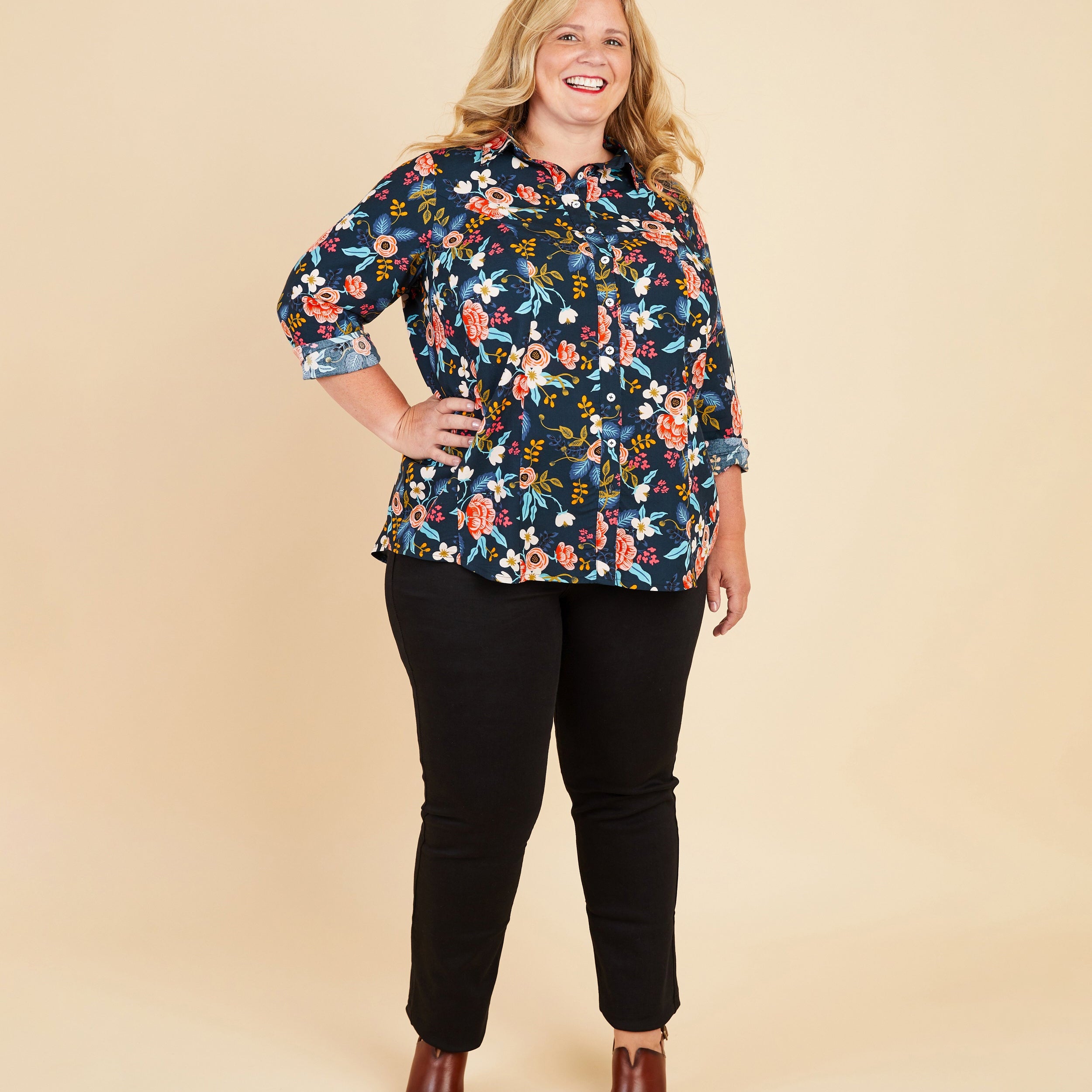 Woman wearing a floral blouse and black pants on a beige background. This button-up shirt is the Harrison Shirt sewing pattern from Cashmerette.