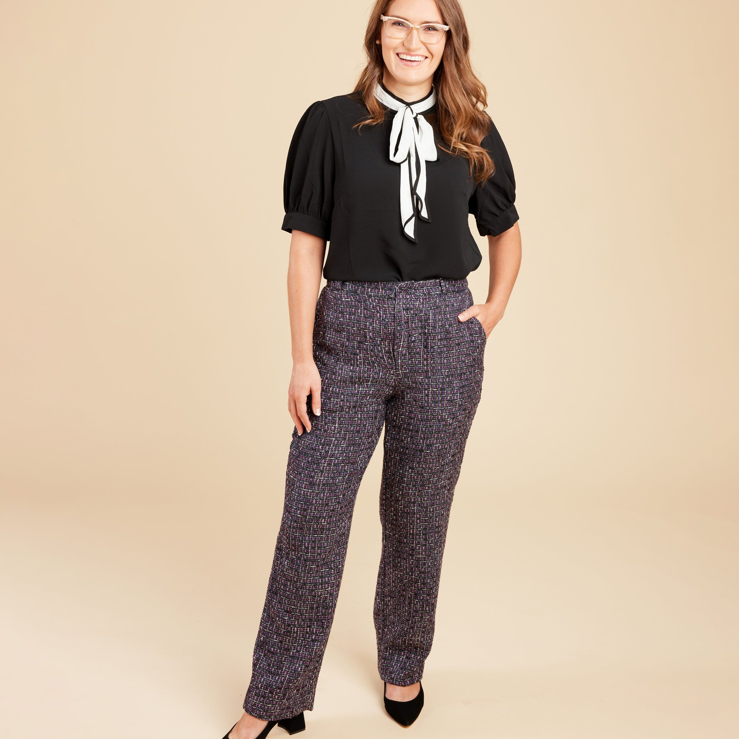 Woman wearing a black blouse with a white bow and patterned pants on a beige background. These pants are the Meriam Trousers sewing pattern from Cashmerette.