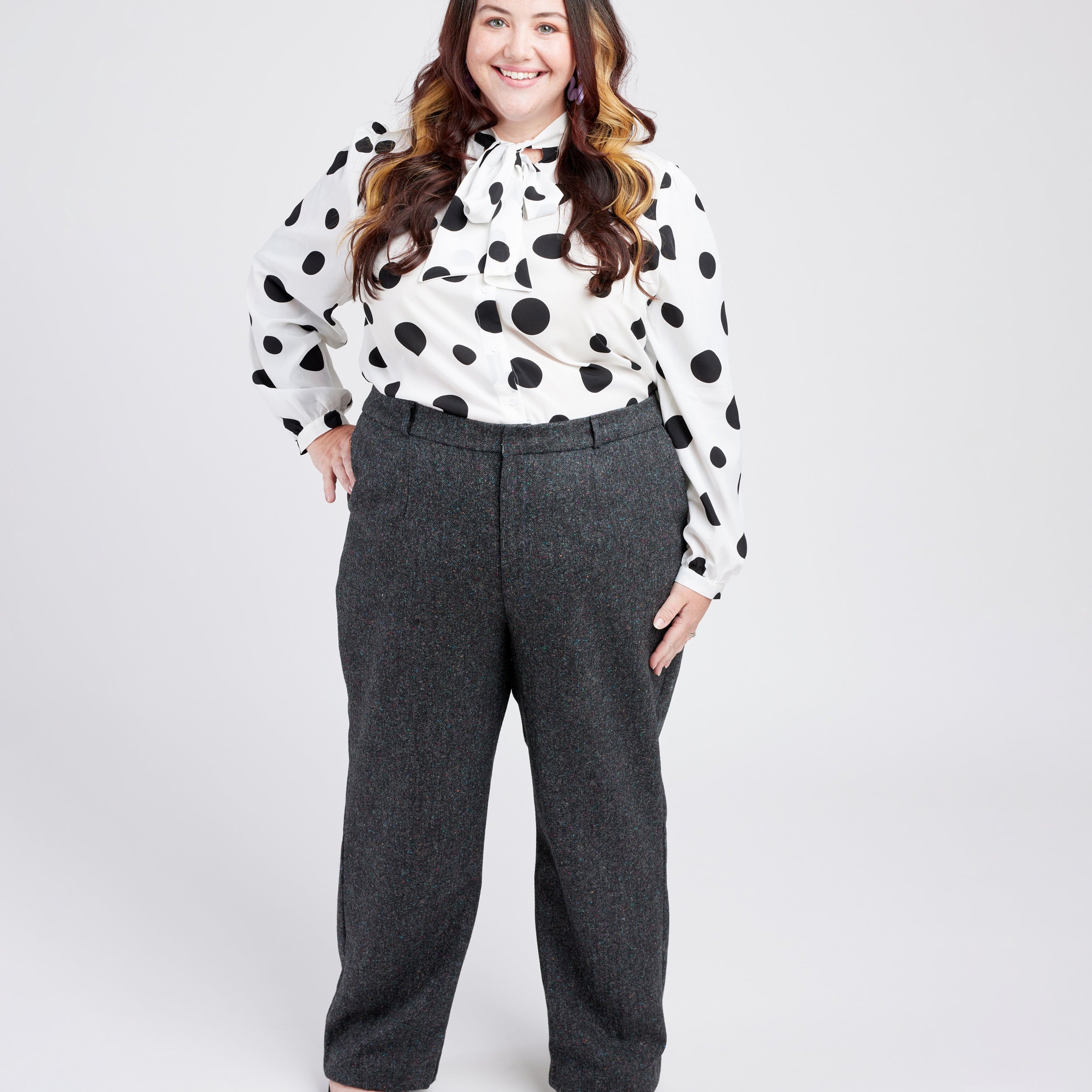 Woman wearing a polka dot blouse and gray pants on a white background. These pants are the Meriam Trousers sewing pattern from Cashmerette.