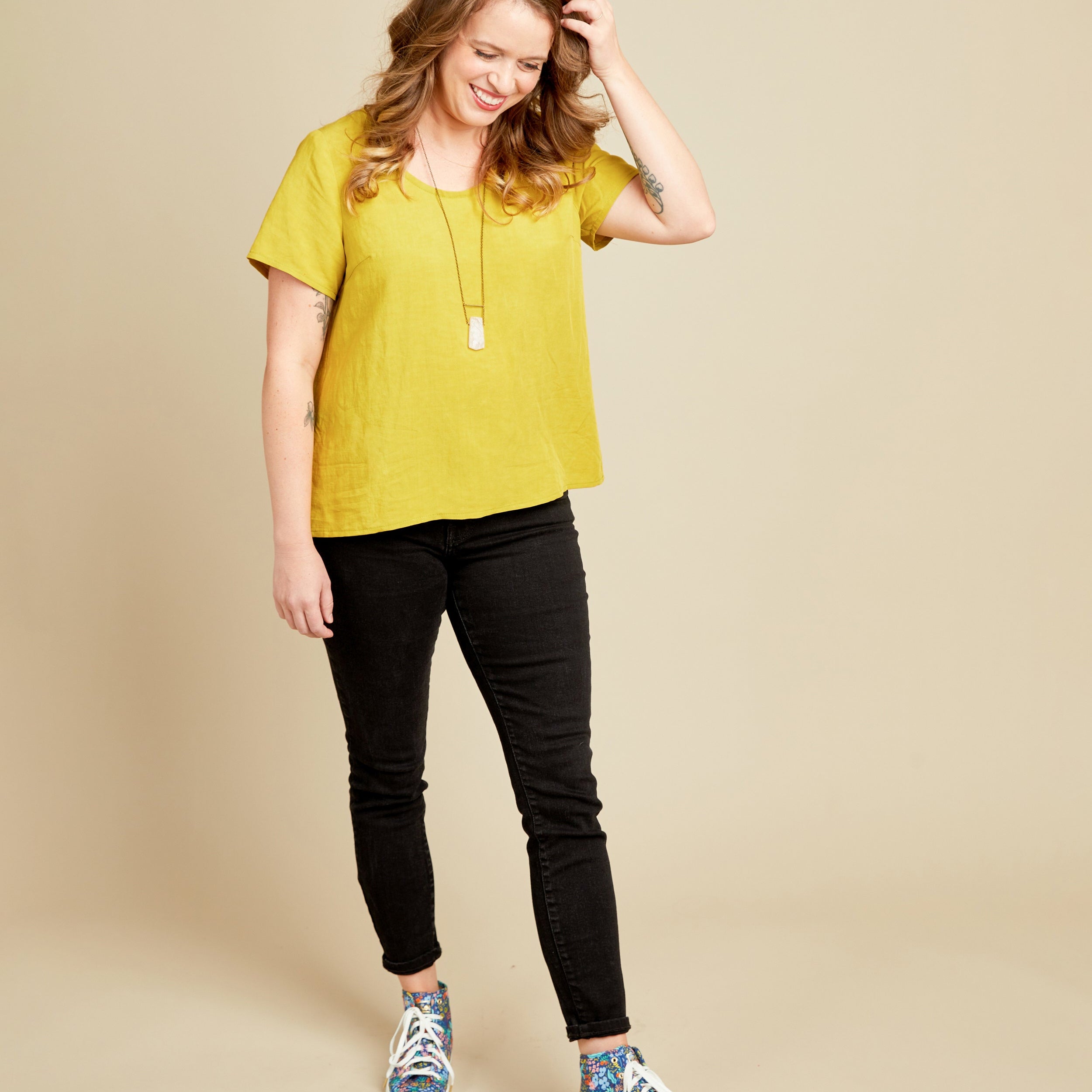 Woman wearing a yellow top and black pants with colorful shoes on a beige background. This top is the Montrose Top sewing pattern from Cashmerette.