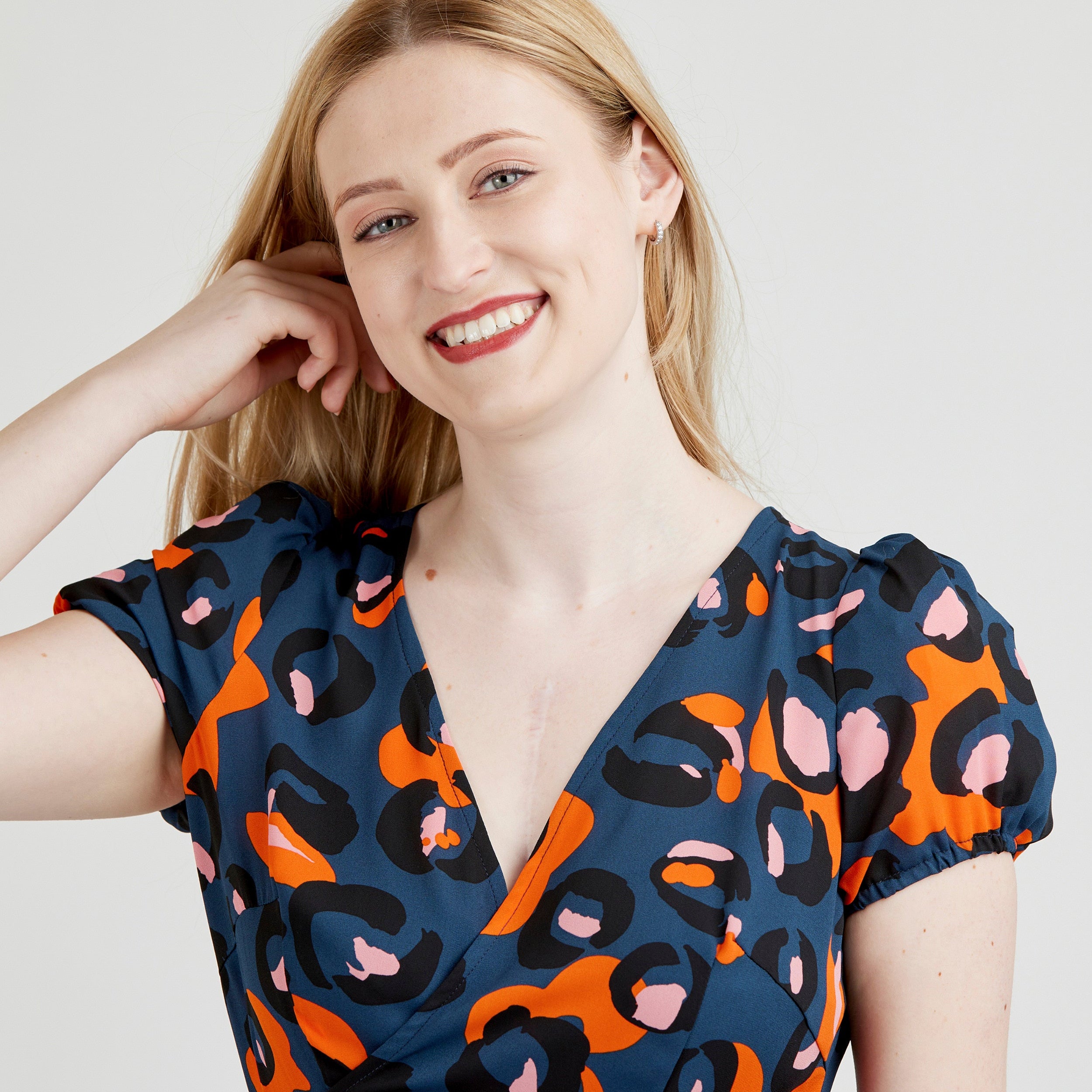 Woman wearing a blue dress with orange and pink abstract pattern on a plain background. This dress is the Roseclair Wrap Dress sewing pattern from Cashmerette.