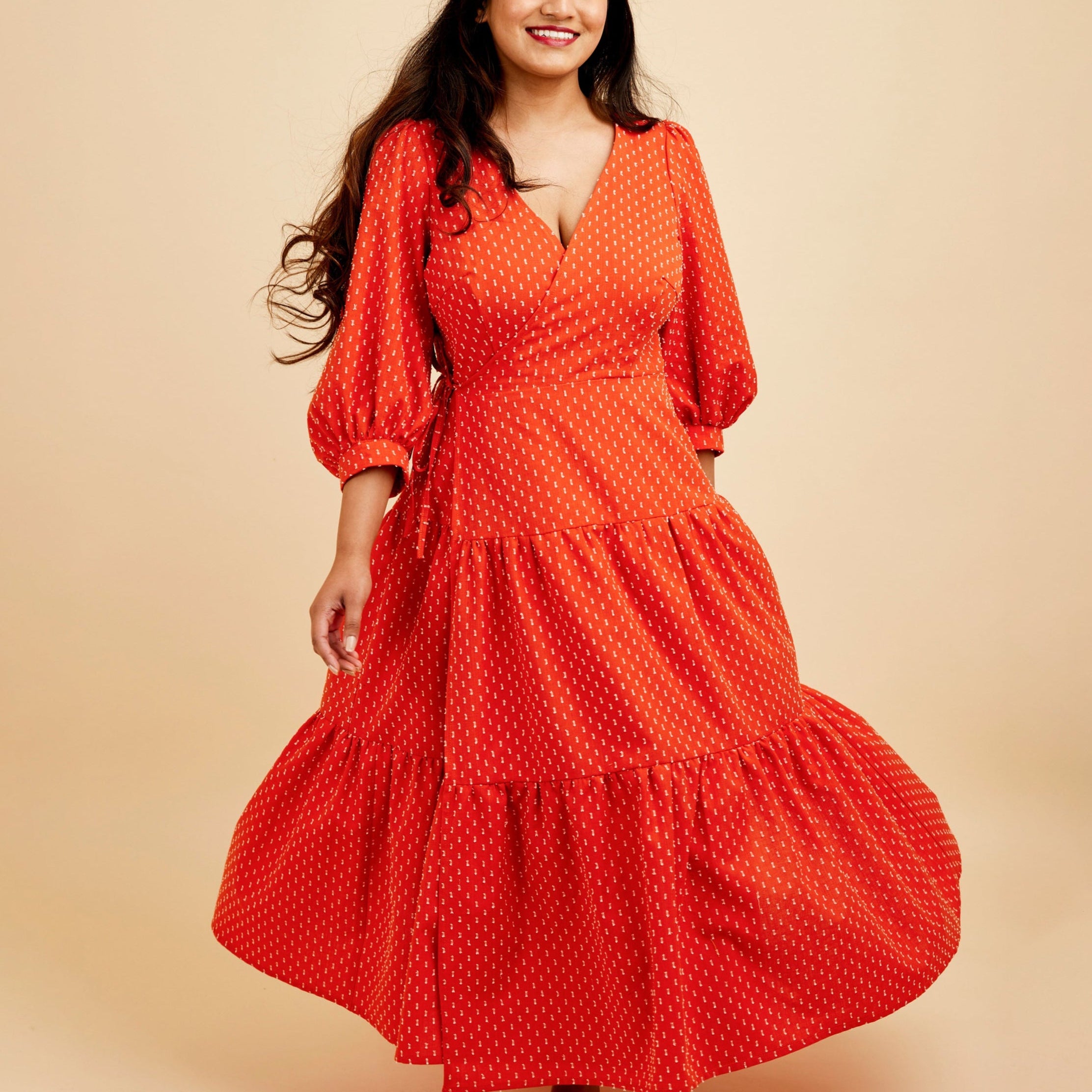 Woman wearing a red polka dot dress on a beige background. This dress is the Roseclair Wrap Dress sewing pattern from Cashmerette.