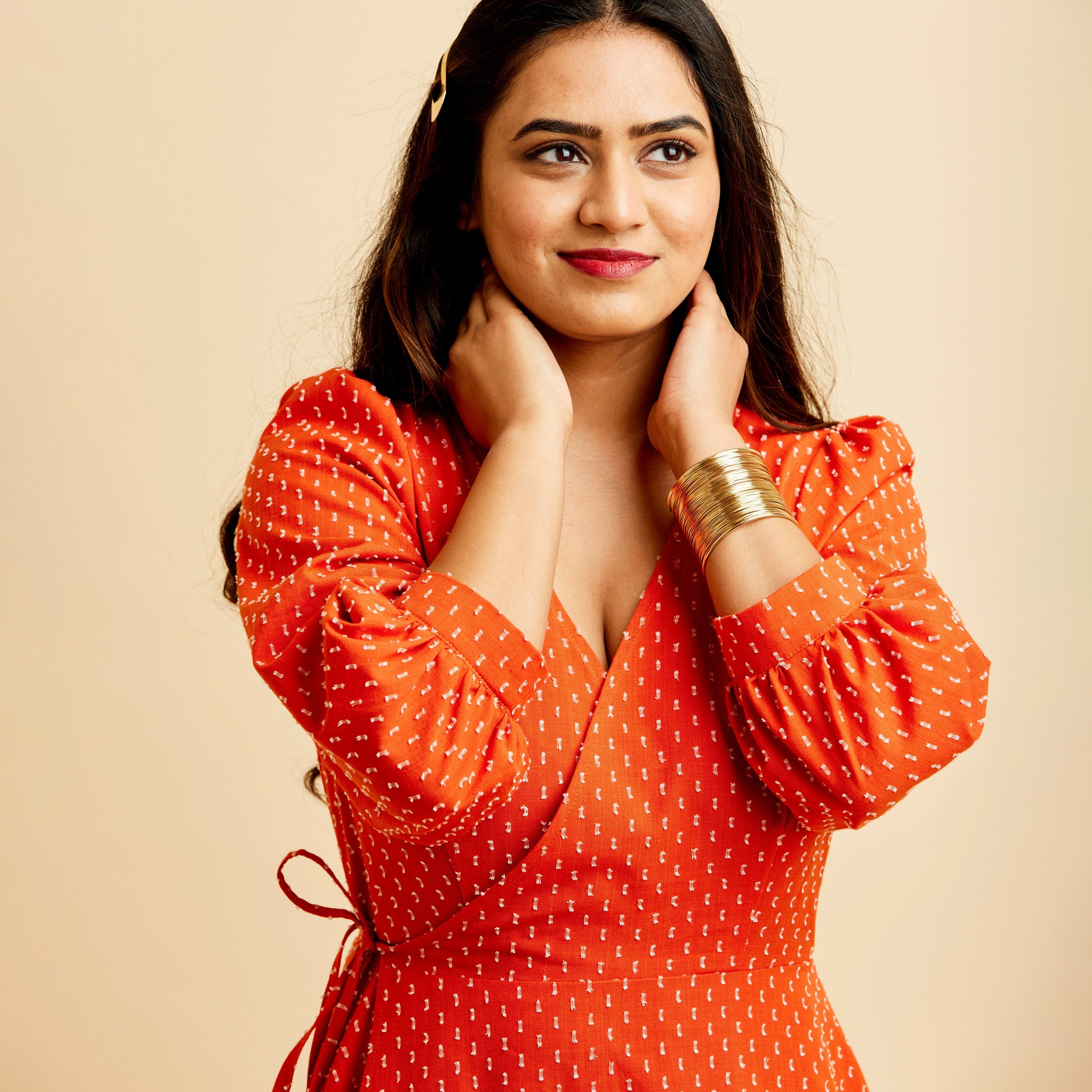 Woman wearing an orange dress with a beige background. This dress is the Roseclair Wrap Dress sewing pattern from Cashmerette.