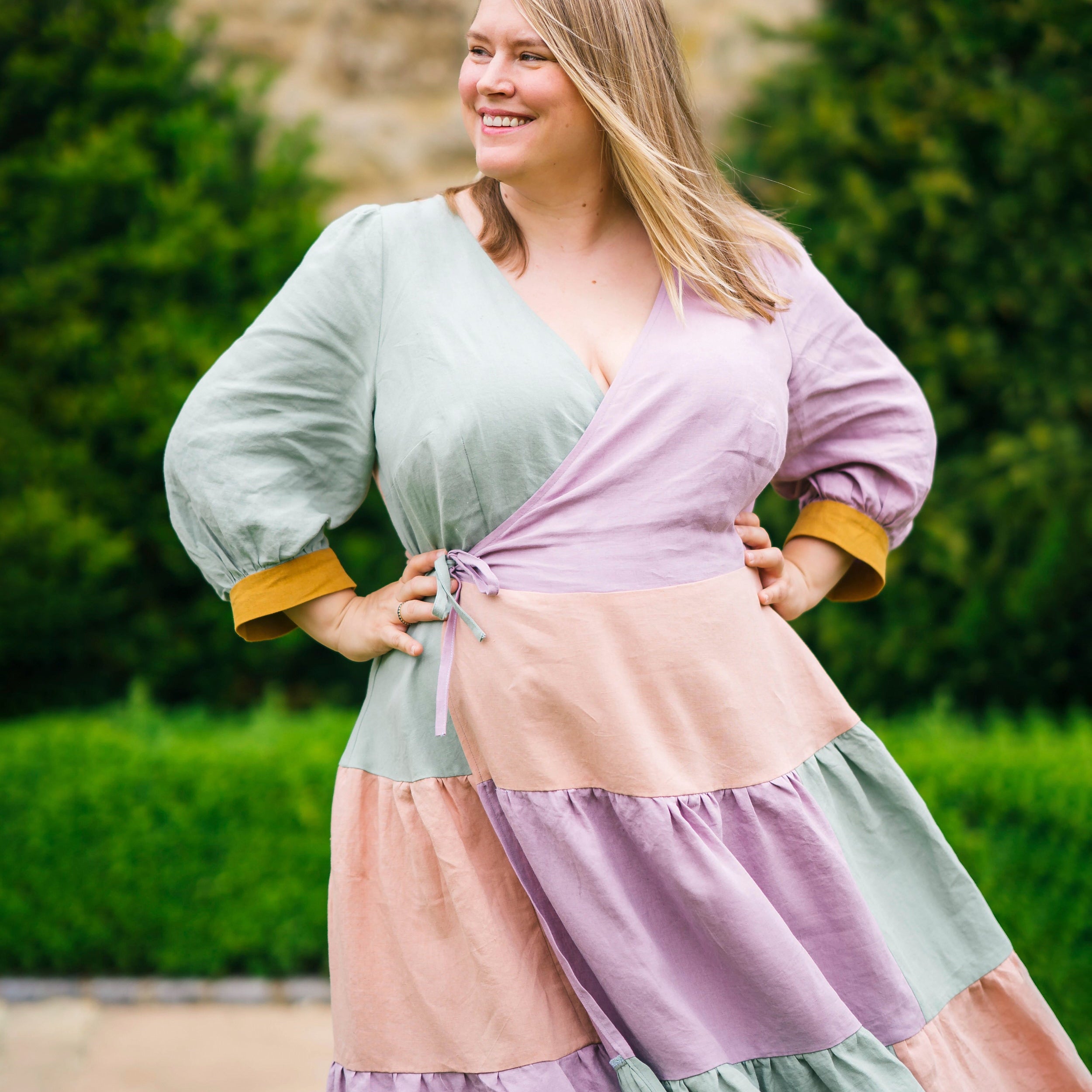 Woman wearing a colorful dress with a green, pink, and purple pattern standing outdoors.This dress is the Roseclair Wrap Dress sewing pattern from Cashmerette.
