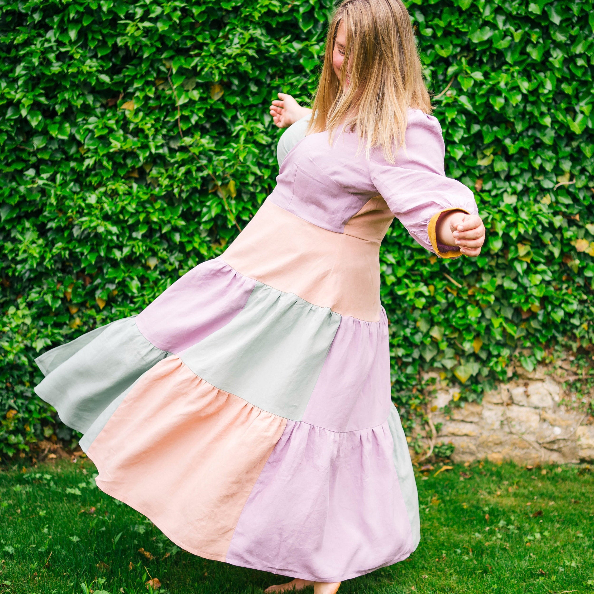 Woman in a pastel-colored dress twirling in front of a green hedge.. This dress is the Roseclair Wrap Dress sewing pattern from Cashmerette.