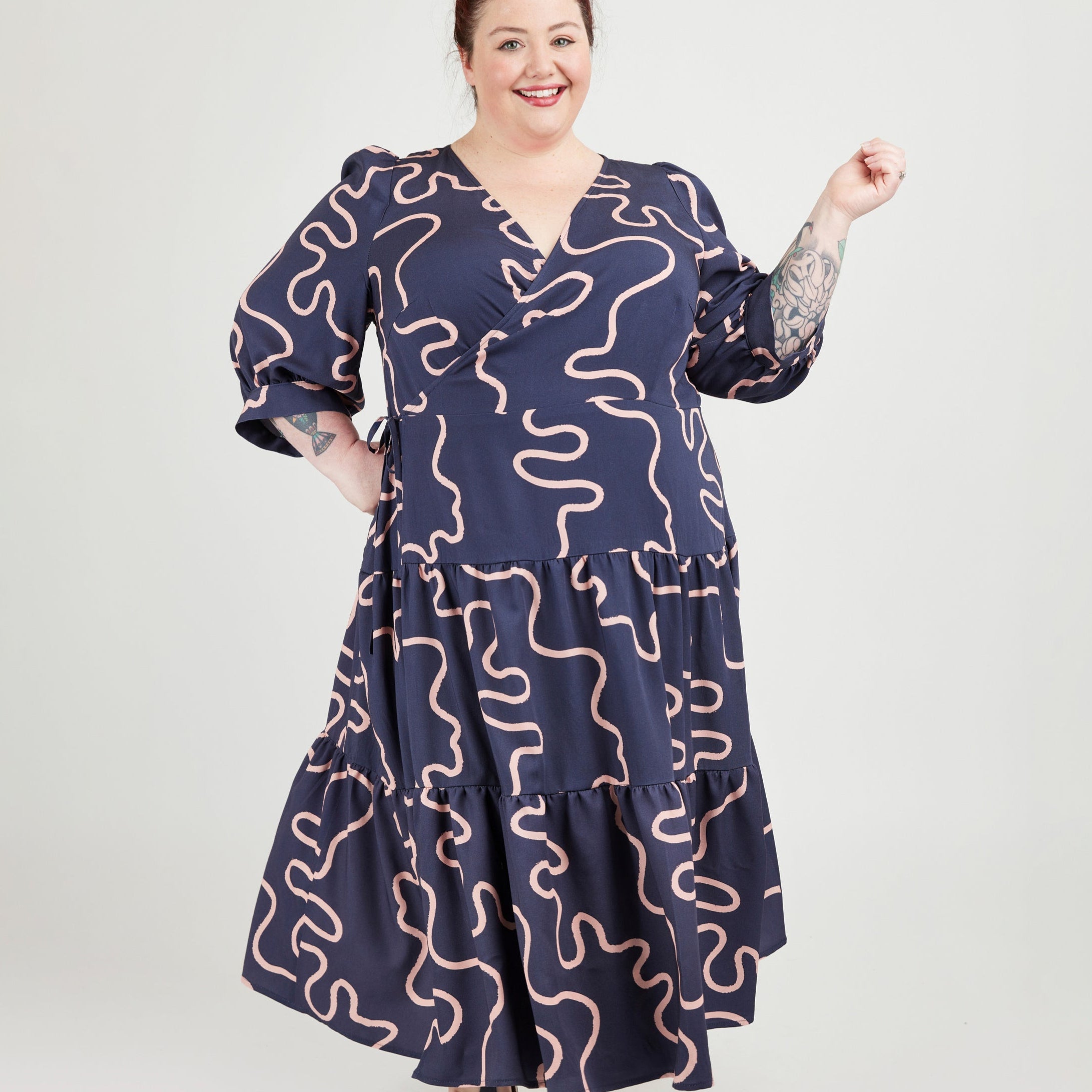 Woman wearing a navy dress with pink wavy patterns on a white background. This dress is the Roseclair Wrap Dress sewing pattern from Cashmerette.