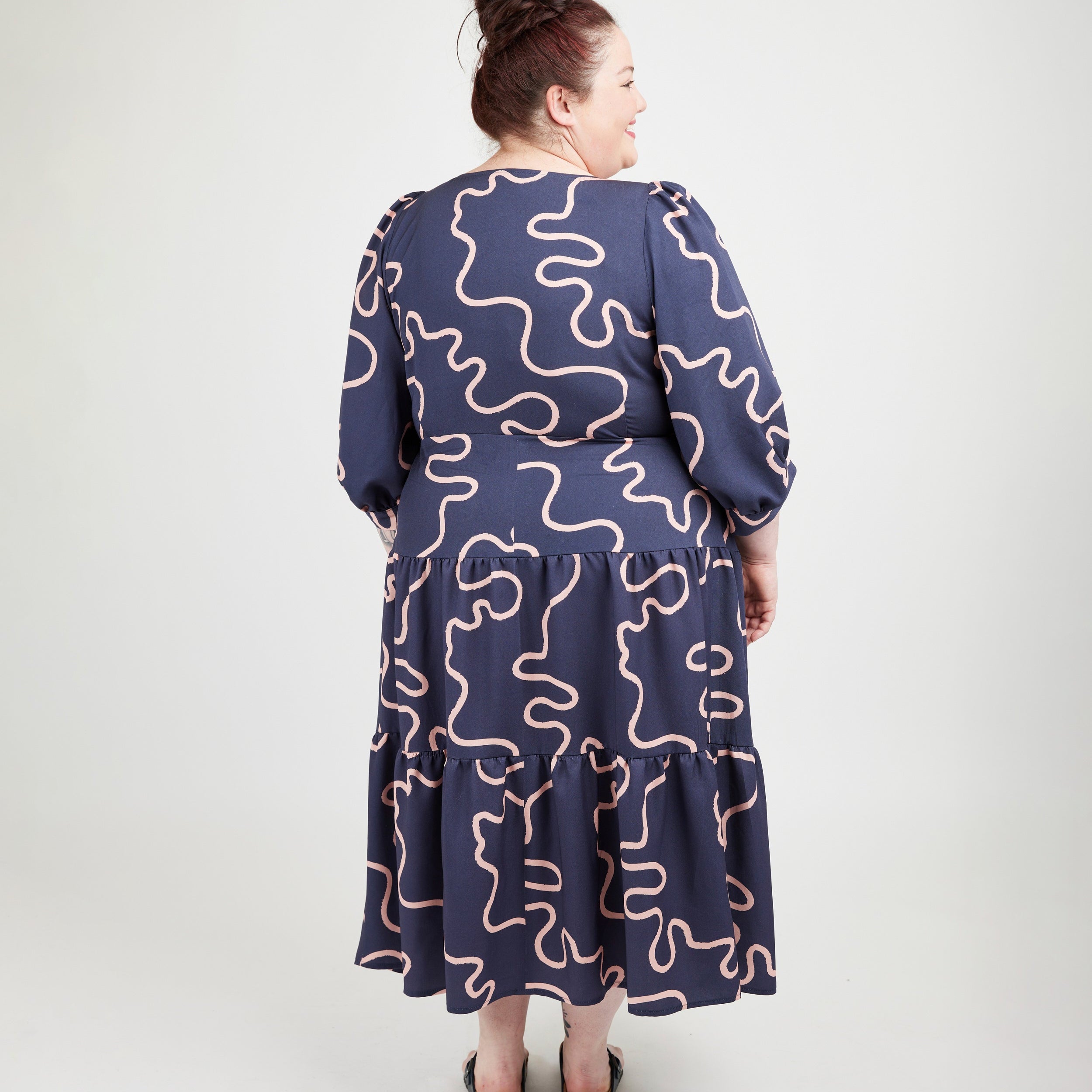 Woman wearing a navy dress with pink wavy patterns on a white background. This dress is the Roseclair Wrap Dress sewing pattern from Cashmerette.