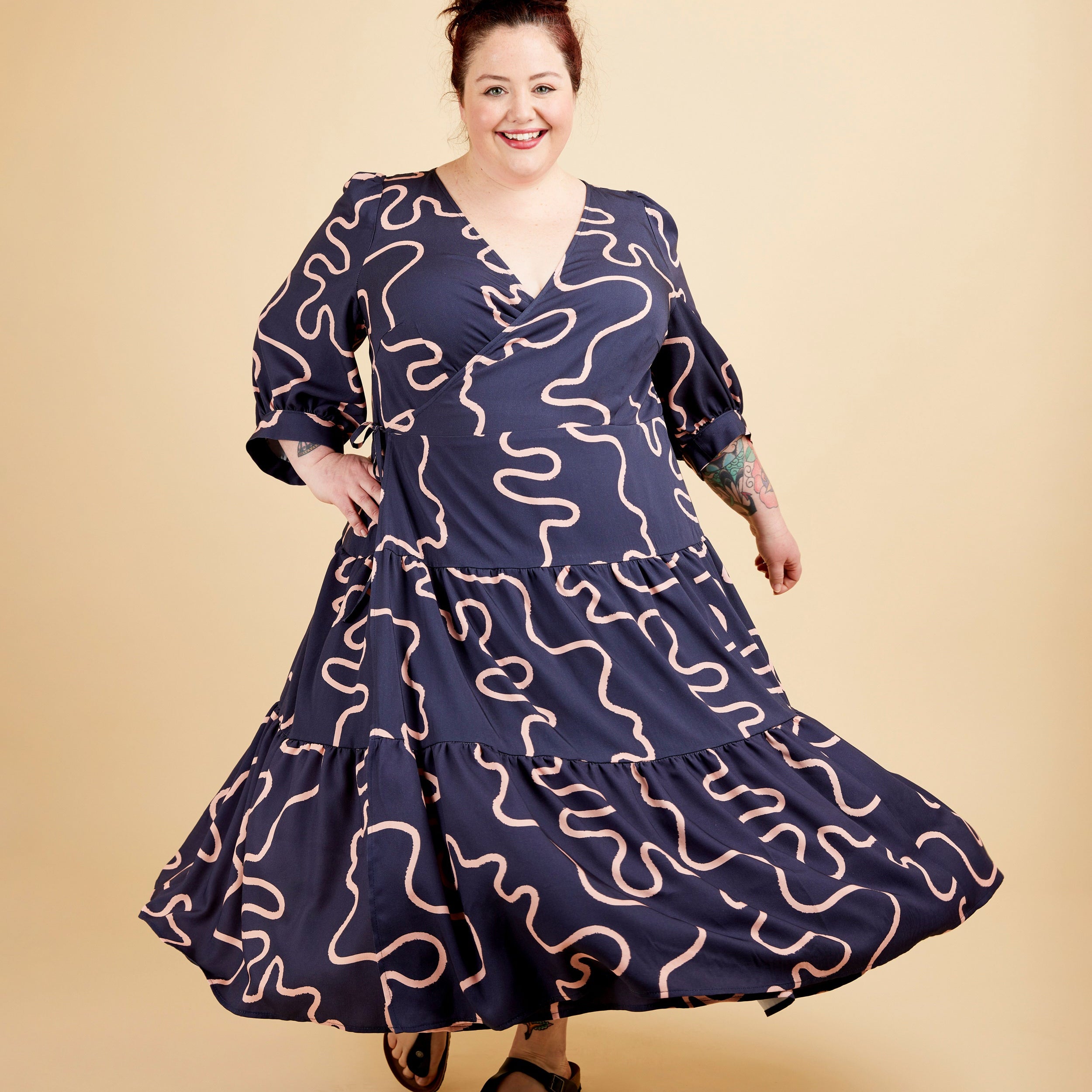 Woman wearing a navy dress with pink patterns on a beige background. This dress is the Roseclair Wrap Dress sewing pattern from Cashmerette.