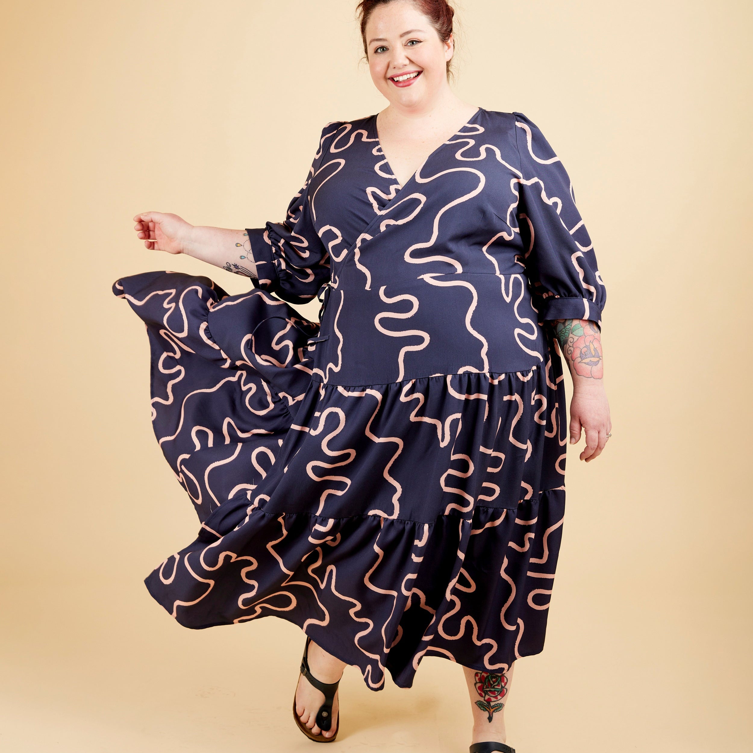 Woman wearing a navy dress with wavy pattern on a beige background. This dress is the Roseclair Wrap Dress sewing pattern from Cashmerette.