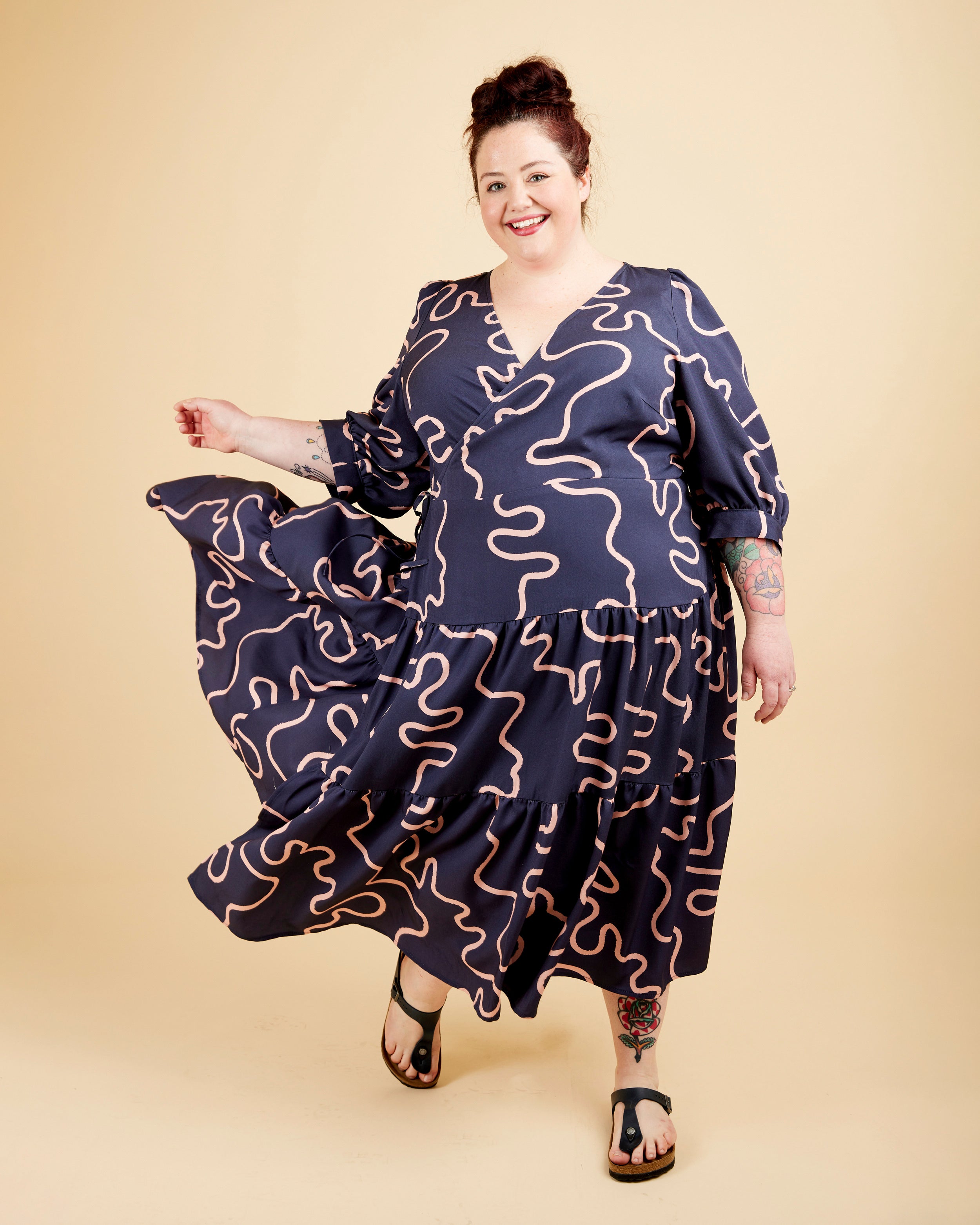 Woman wearing a navy dress with wavy pattern on a beige background. This dress is the Roseclair Wrap Dress sewing pattern from Cashmerette.