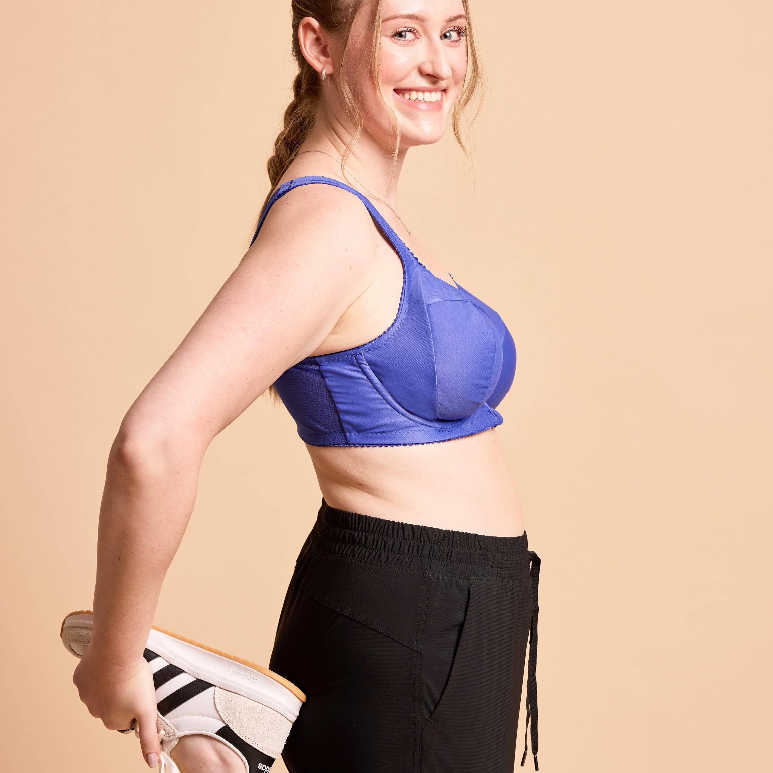 Woman wearing a blue sports bra and black leggings, holding white sneakers against a beige background. This is the Cashmerette Somerville Sports Bra sewing pattern for big boobs.