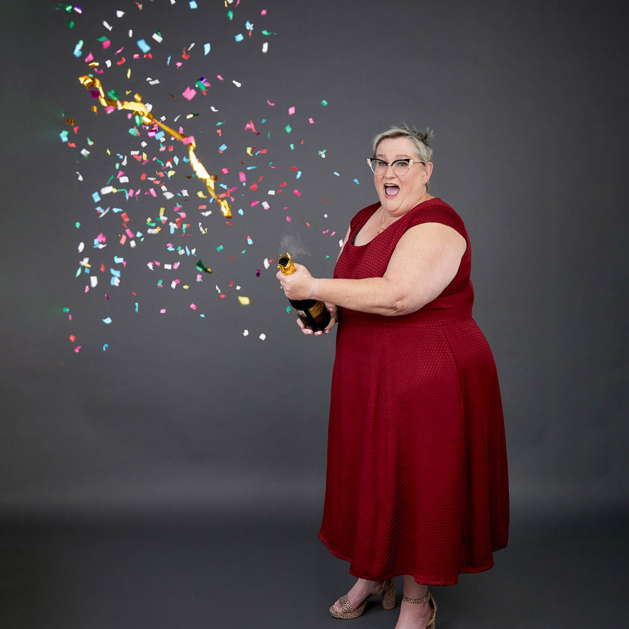 Woman in a red dress holding a bottle with confetti falling around her on a dark background. This dress is the Upton Dress & Skirt sewing pattern from Cashmerette.