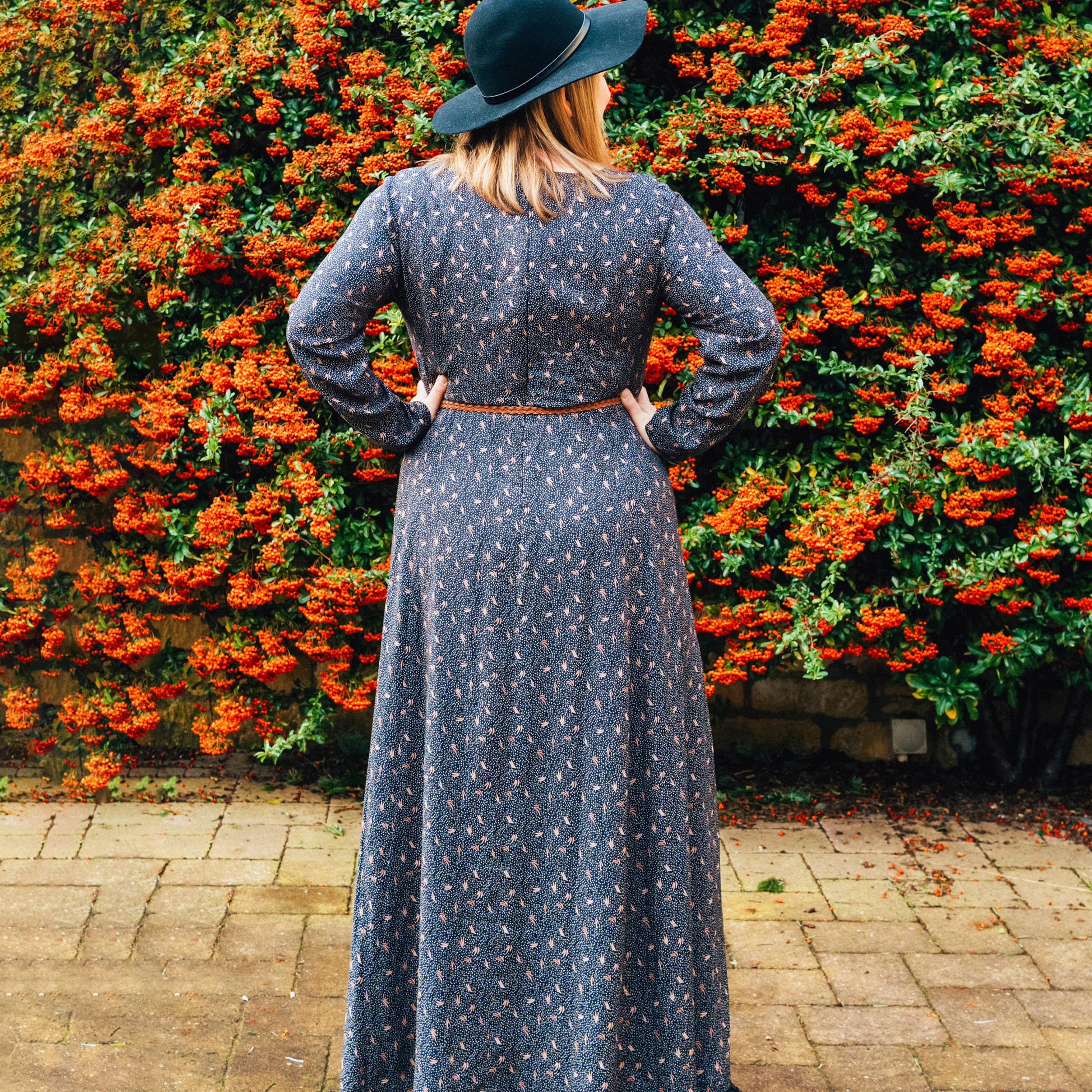 Woman in a long dark dress and black hat standing in front of a wall of orange flowers.. This dress is the Upton Dress & Skirt sewing pattern from Cashmerette.