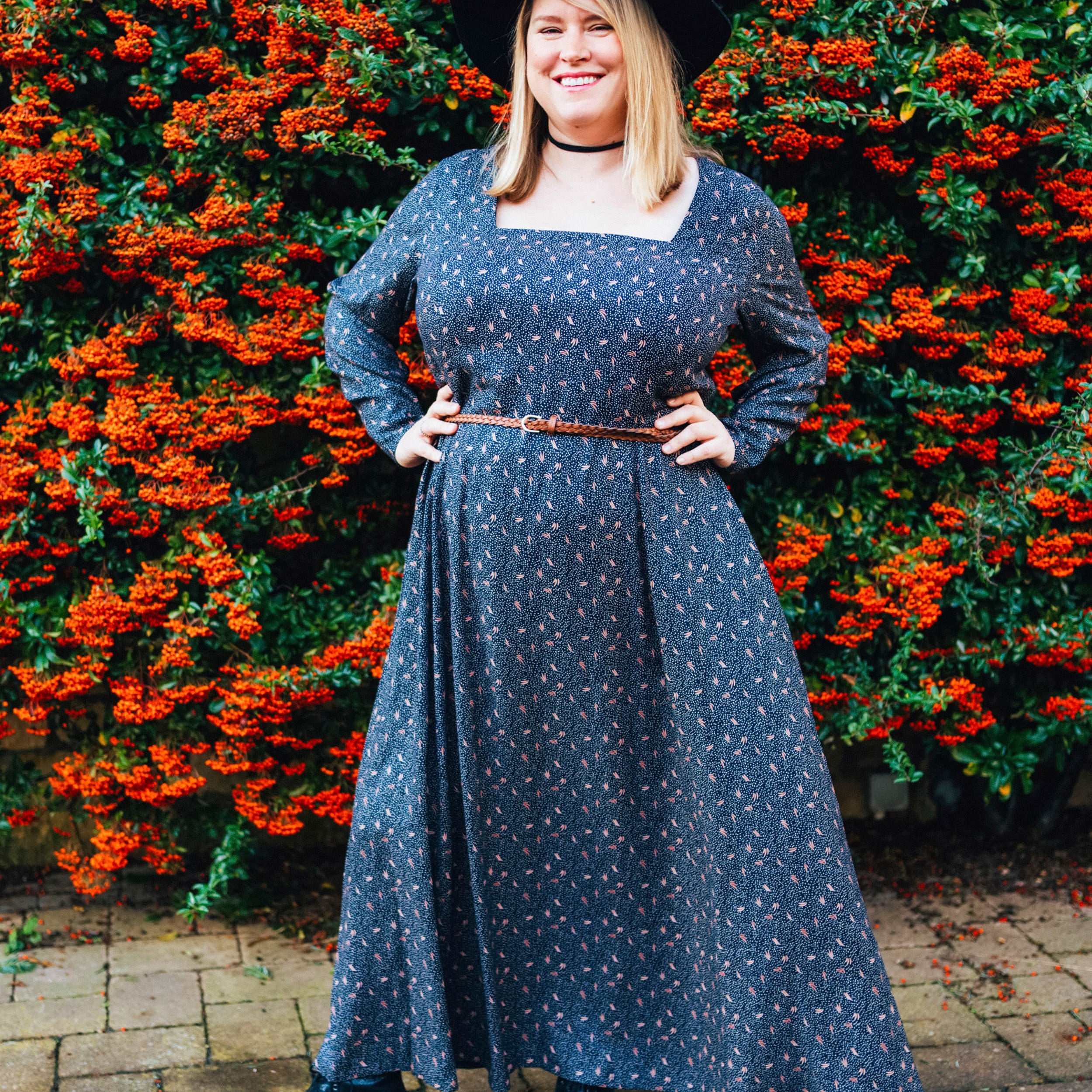 Woman in a blue dress standing in front of a hedge with red flowers. This dress is the Upton Dress & Skirt sewing pattern from Cashmerette.