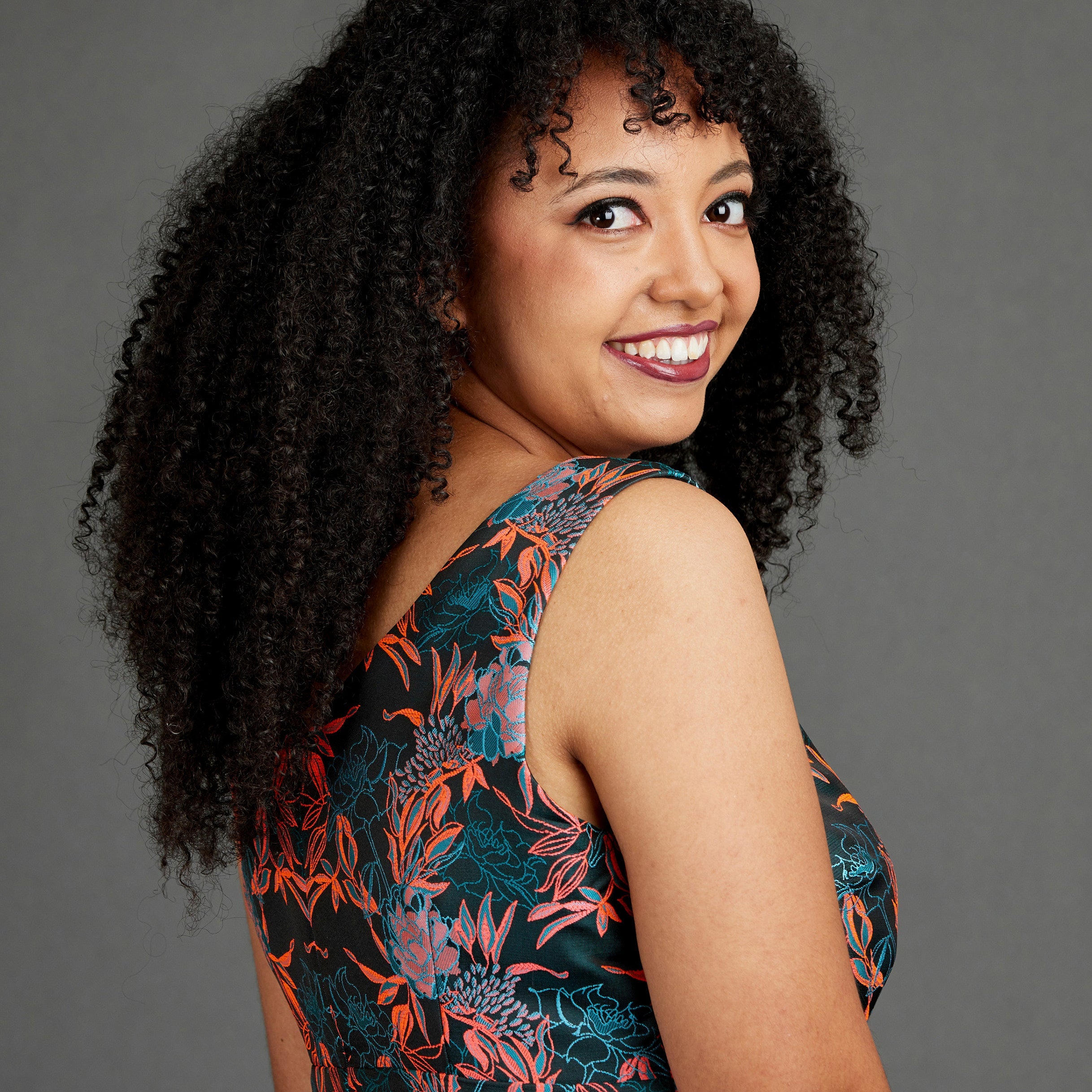 Woman with curly hair wearing a floral dress against a gray background. This dress is the Upton Dress & Skirt sewing pattern from Cashmerette.