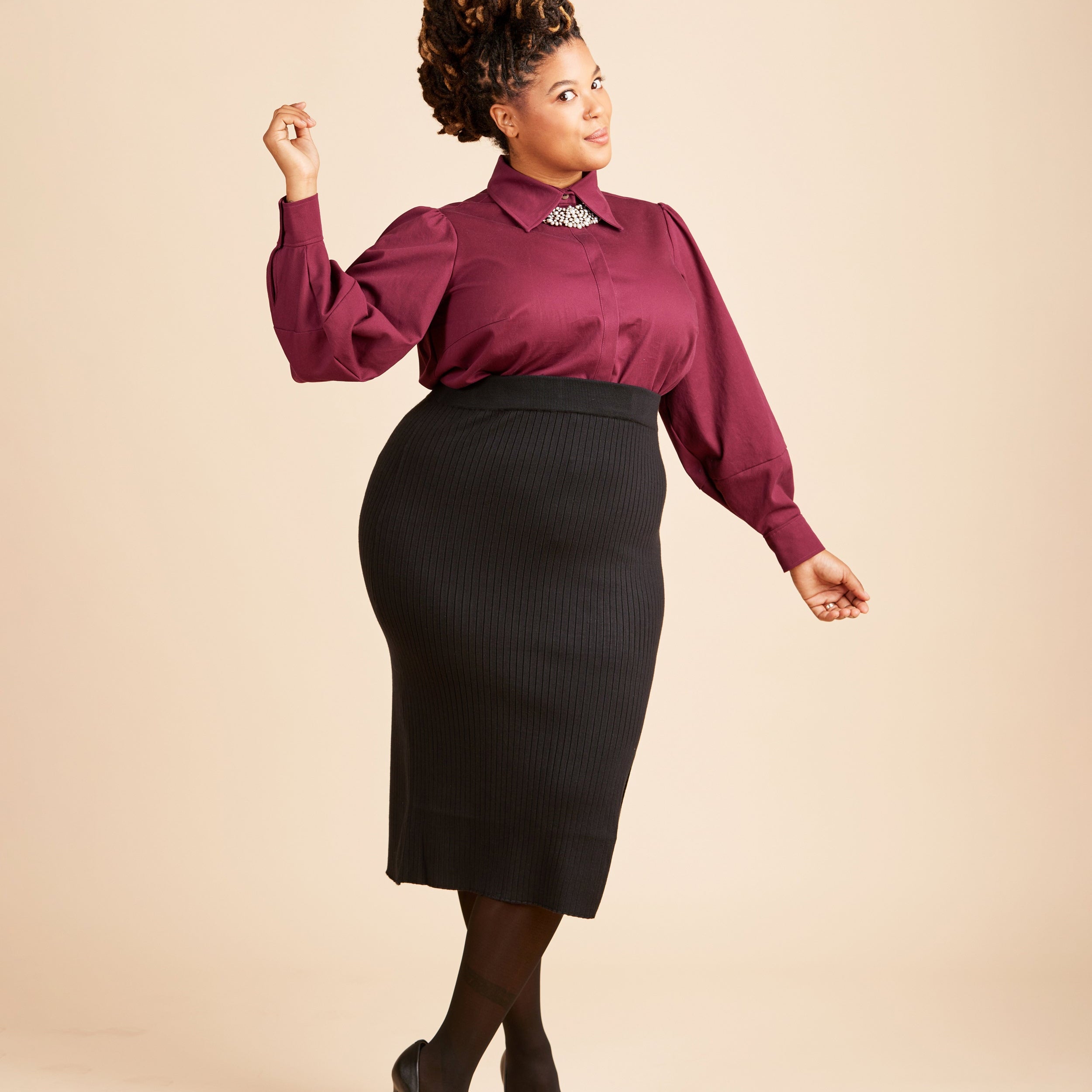 Woman wearing a burgundy blouse and black skirt on a beige background. This top is the Vernon Shirt sewing pattern from Cashmerette.