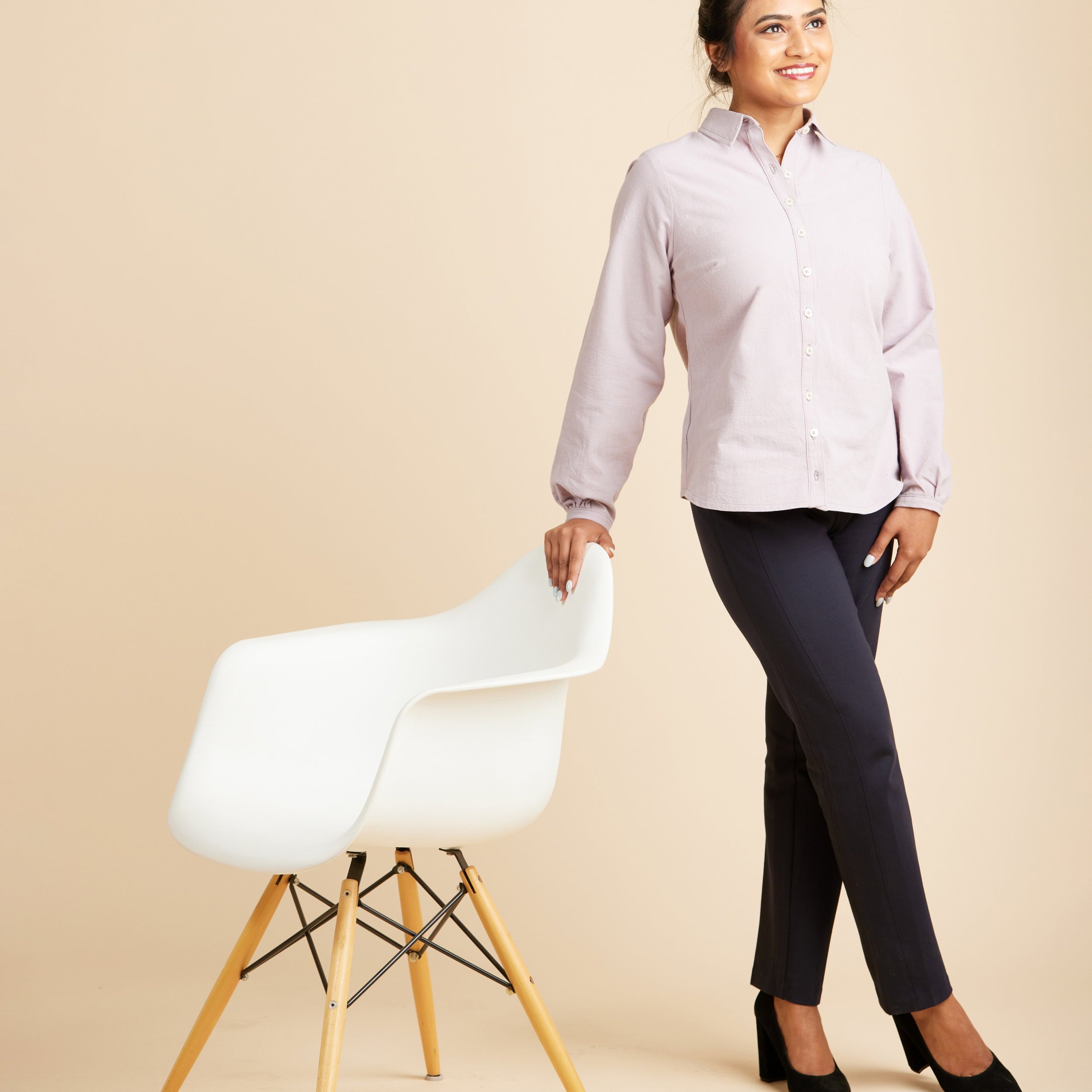 Woman in a light purple shirt and dark pants standing next to a white chair on a beige background. This top is the Vernon Shirt sewing pattern from Cashmerette.