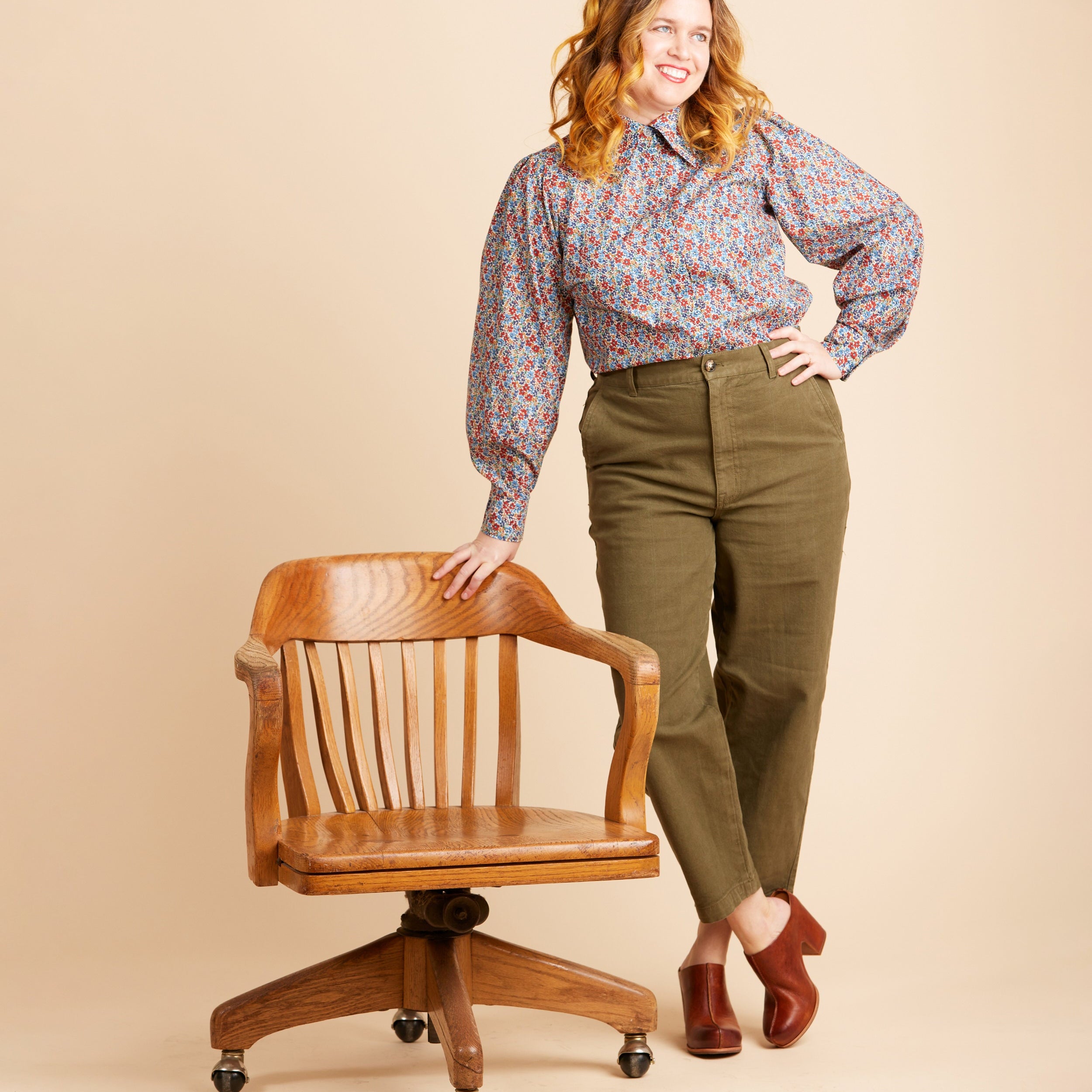 Woman in a patterned blouse and green pants standing next to a wooden chair on a beige background. This top is the Vernon Shirt sewing pattern from Cashmerette.