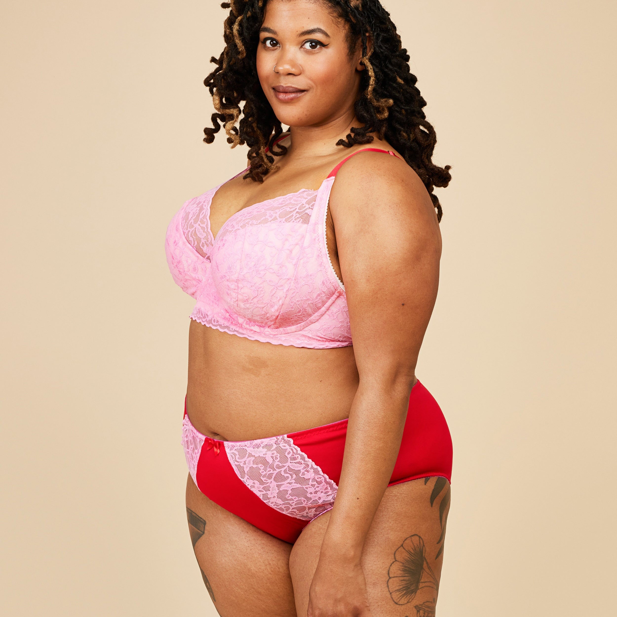 Woman wearing a pink lace bralette and red high-waisted underwear on a beige background. This bra is the Willowdale Bra sewing pattern from Cashmerette.