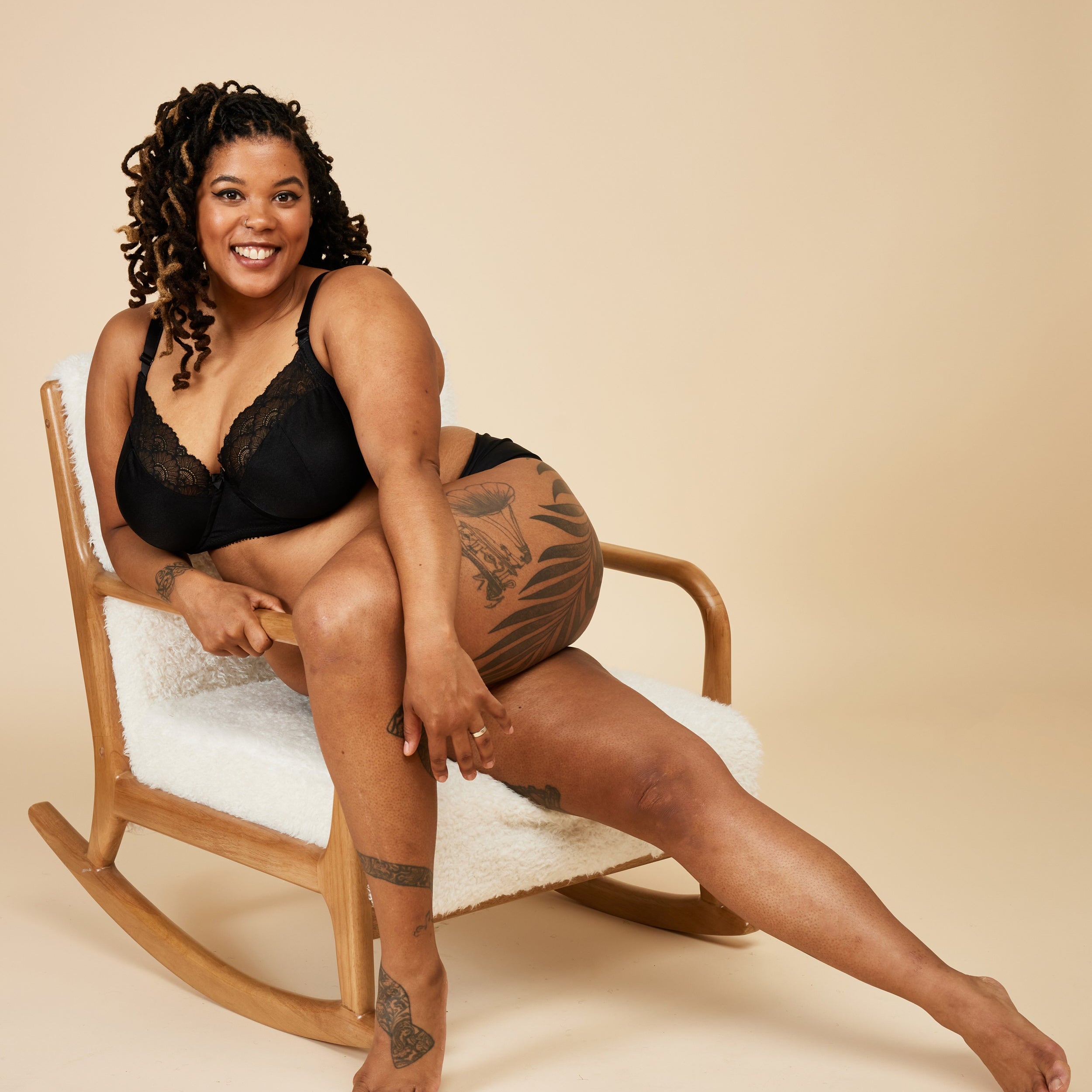 Woman sitting on a rocking chair wearing black lingerie against a beige background. This bra is the Willowdale Bra sewing pattern from Cashmerette.
