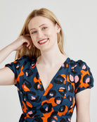 Woman wearing a blue dress with orange and pink abstract pattern on a plain background. This dress is the Roseclair Wrap Dress sewing pattern from Cashmerette.