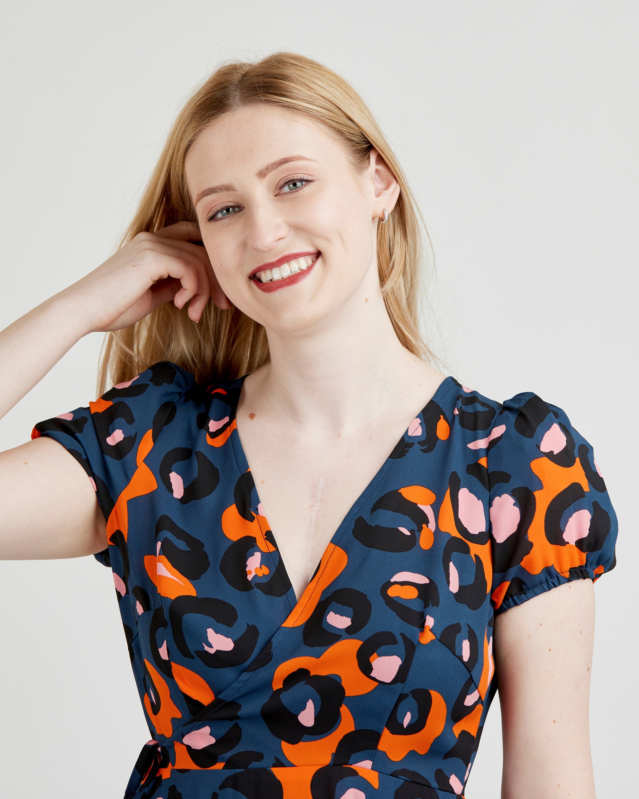 Woman wearing a blue dress with orange and pink abstract pattern on a plain background. This dress is the Roseclair Wrap Dress sewing pattern from Cashmerette.