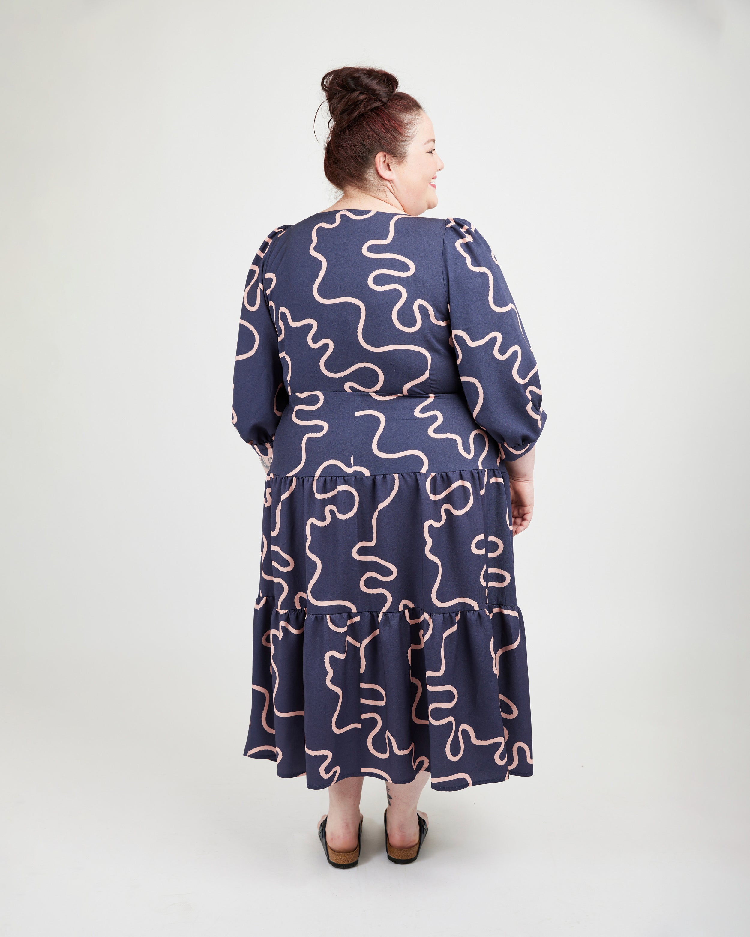 Woman wearing a navy dress with pink wavy patterns on a white background. This dress is the Roseclair Wrap Dress sewing pattern from Cashmerette.